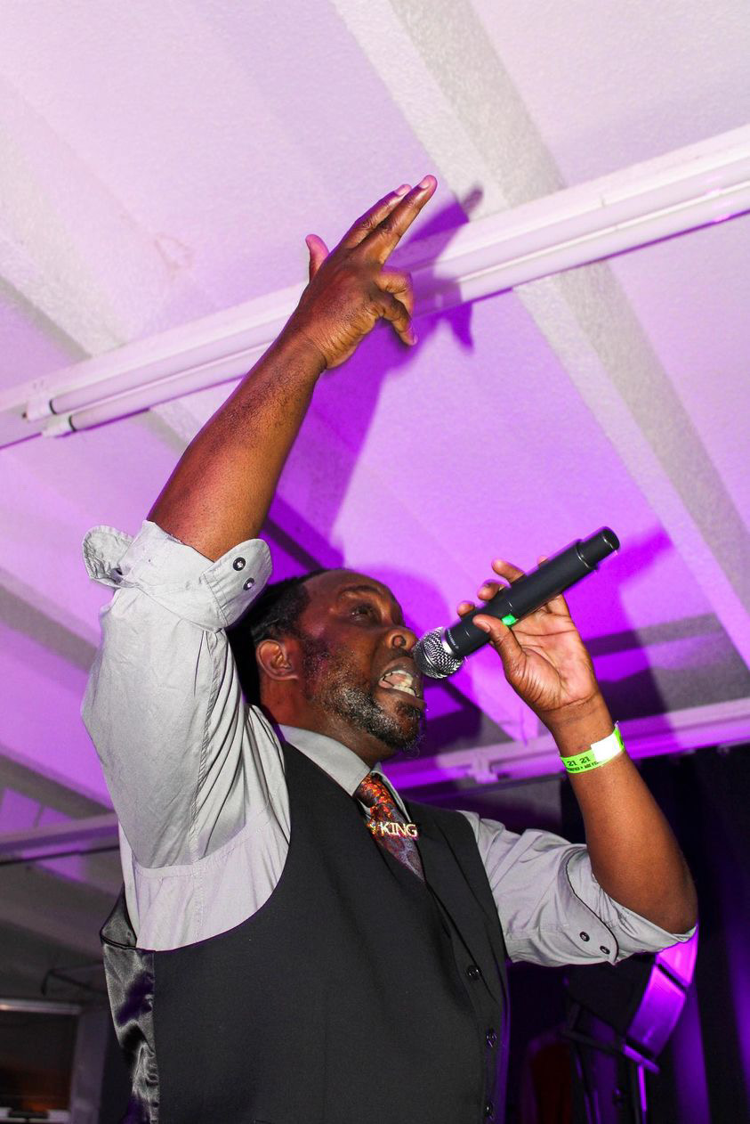 Man singing into microphone with arm raised, wearing a vest and shirt, in a purple-lit setting.