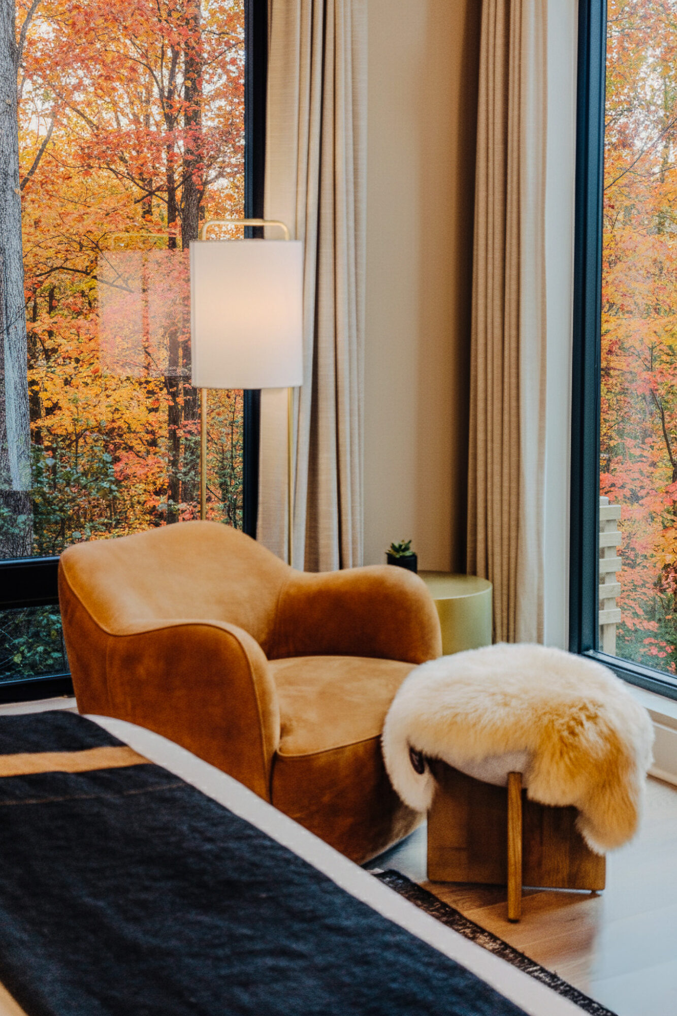 Interior with a beige armchair, a white lamp, and large windows showing autumn trees outside.