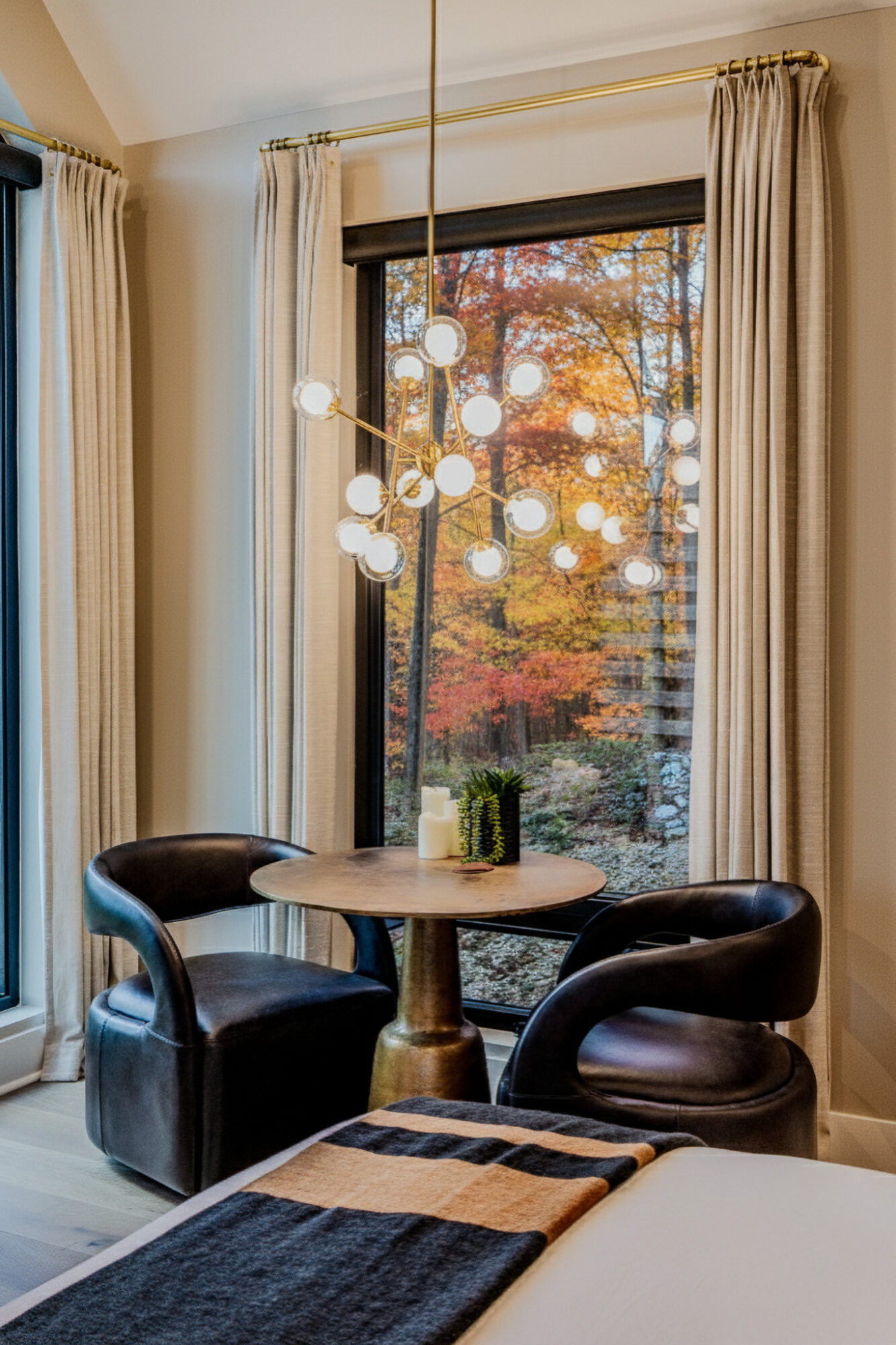 Round table with two black chairs near a window with beige curtains and autumn trees outside.