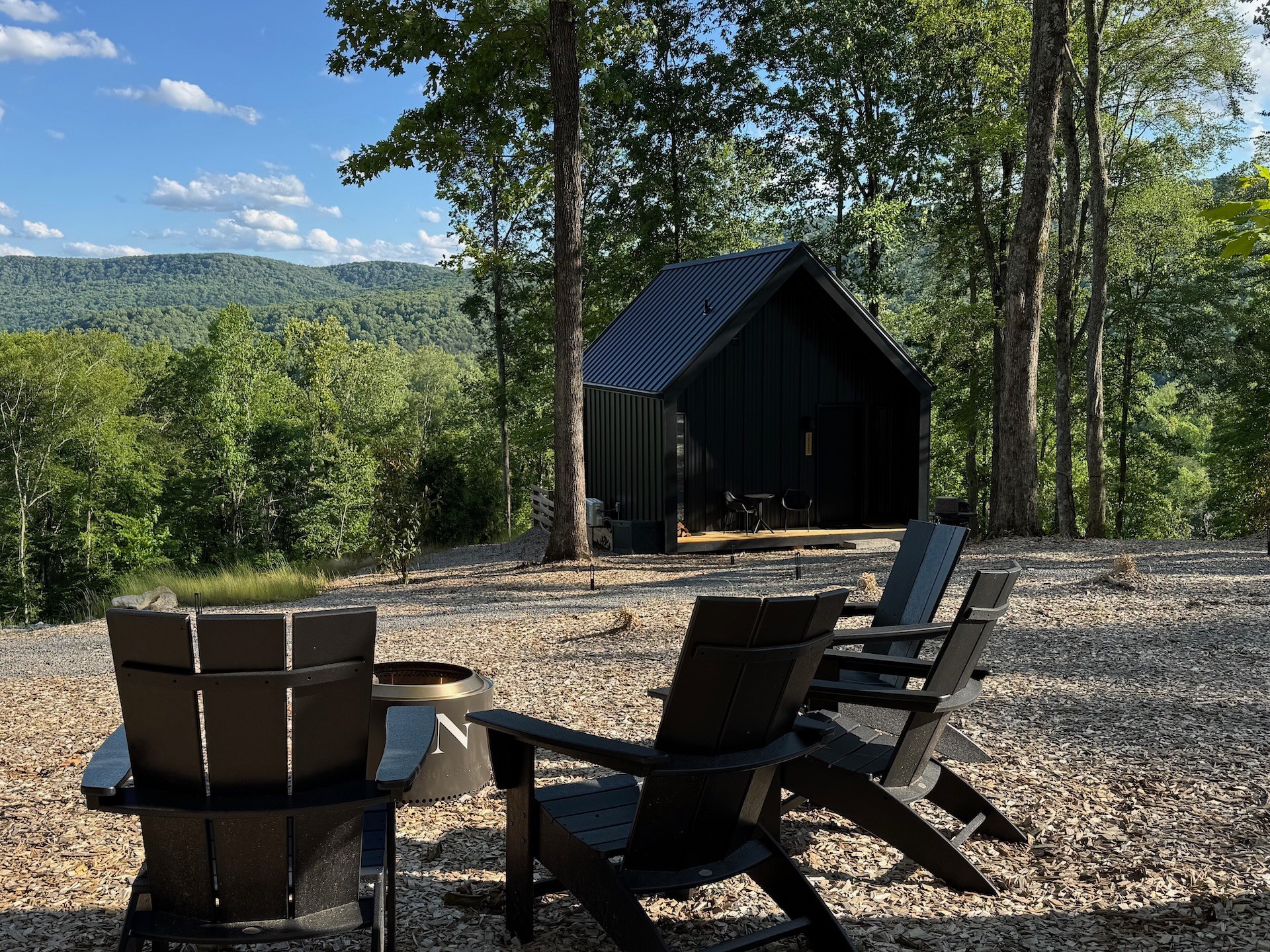 Black wooden shed among trees with outdoor chairs and a table in a natural setting, hills in the background.