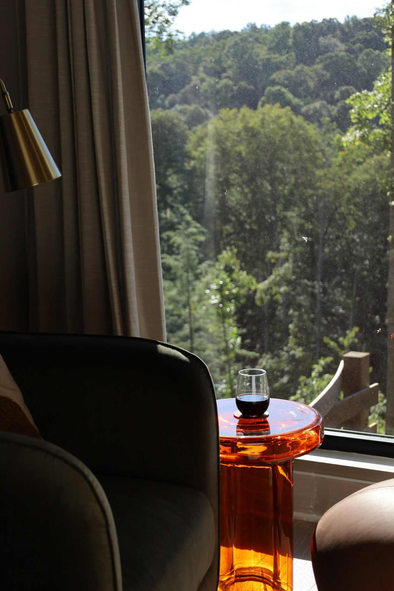 Indoor scene with a window showing trees and greenery outside, a black chair, and an orange glass side table with a glass of water.
