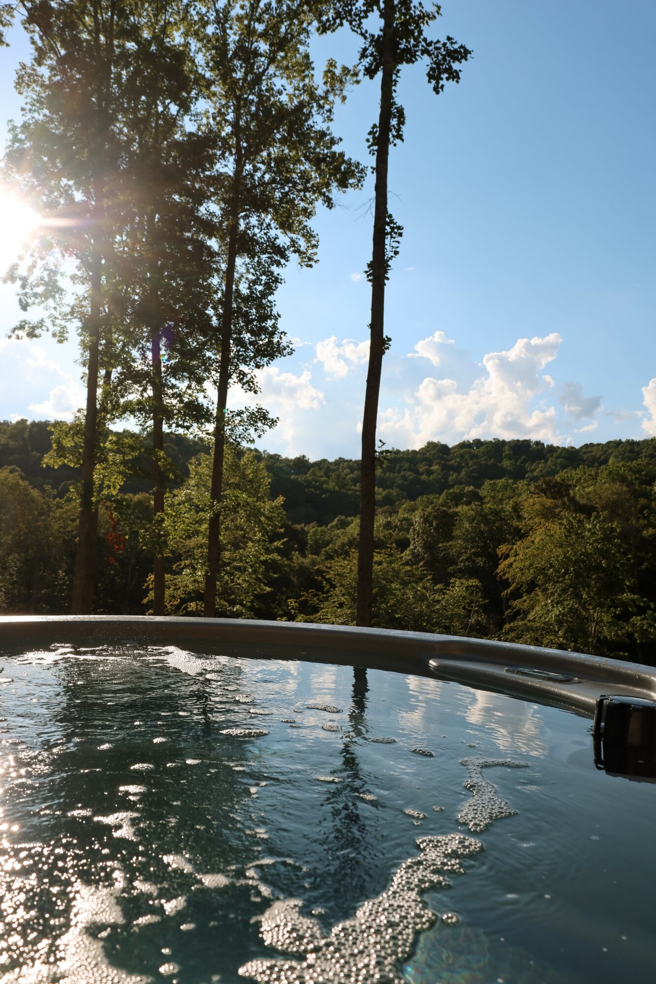 View of trees and sky with sunlight, over a hot tub with bubbling water, outdoors during daytime.