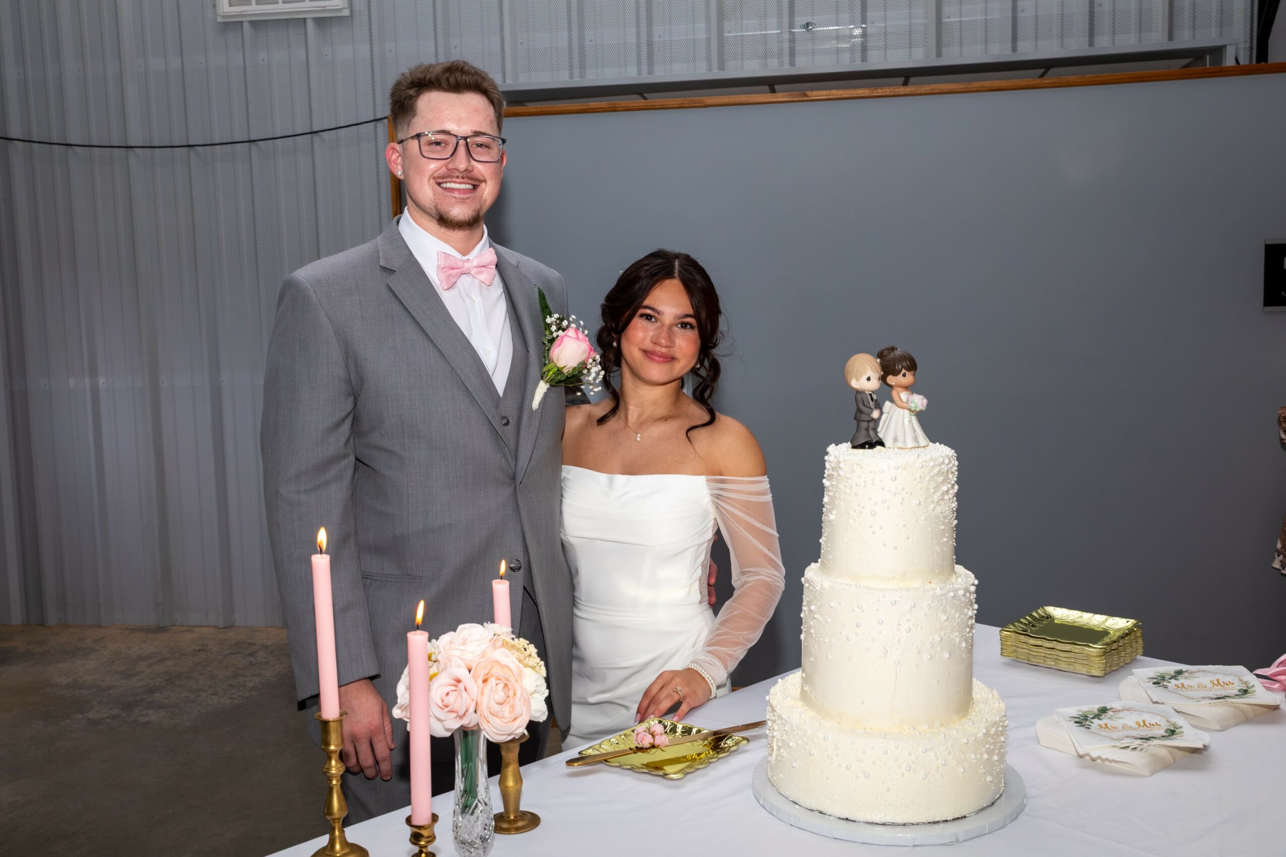 A bride and groom stand together indoors with a three-tiered wedding cake decorated with a bride and groom topper, candles, and flowers.