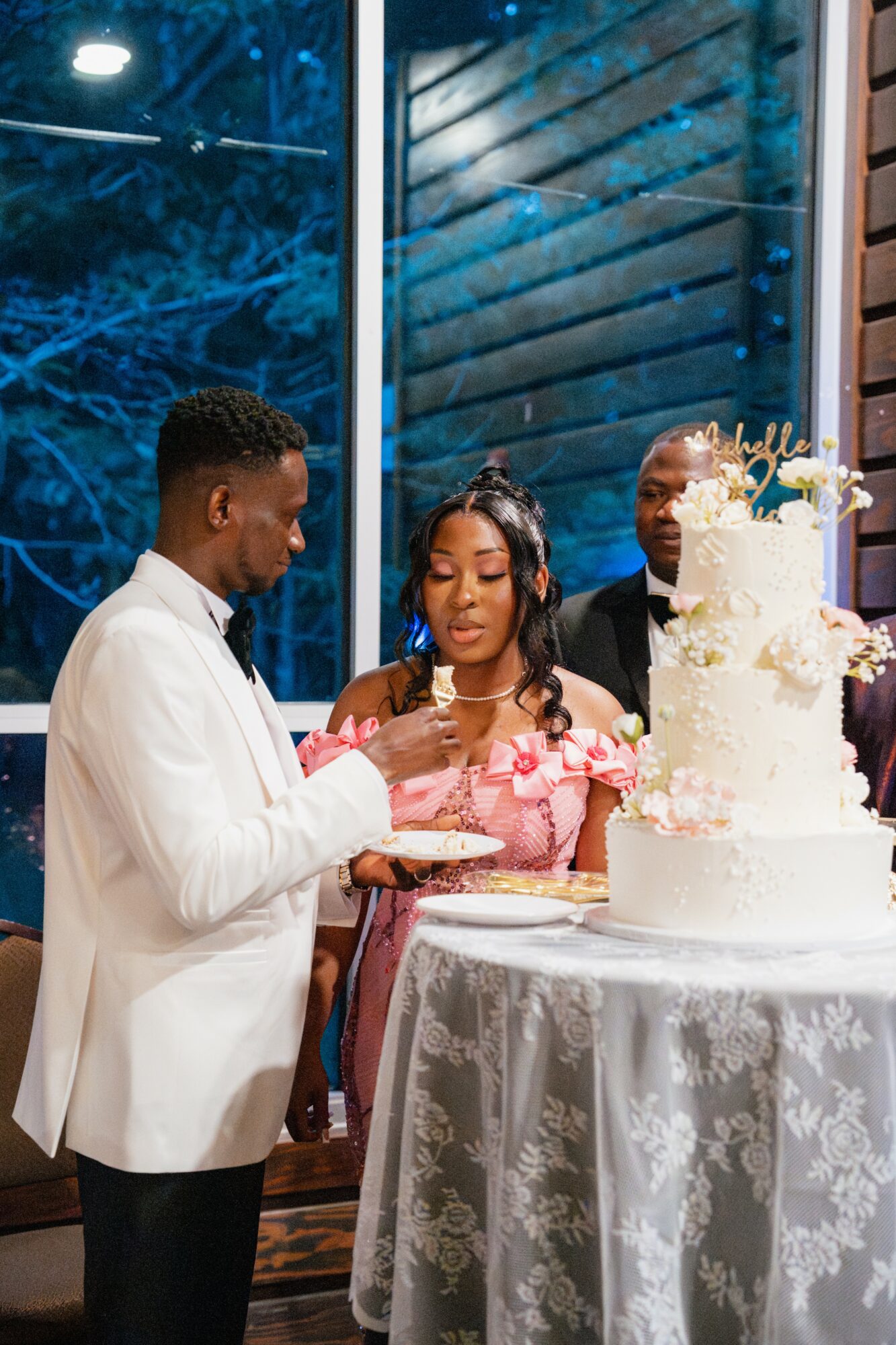 Three people celebrating a wedding with a large white cake, indoors near a window, at night.