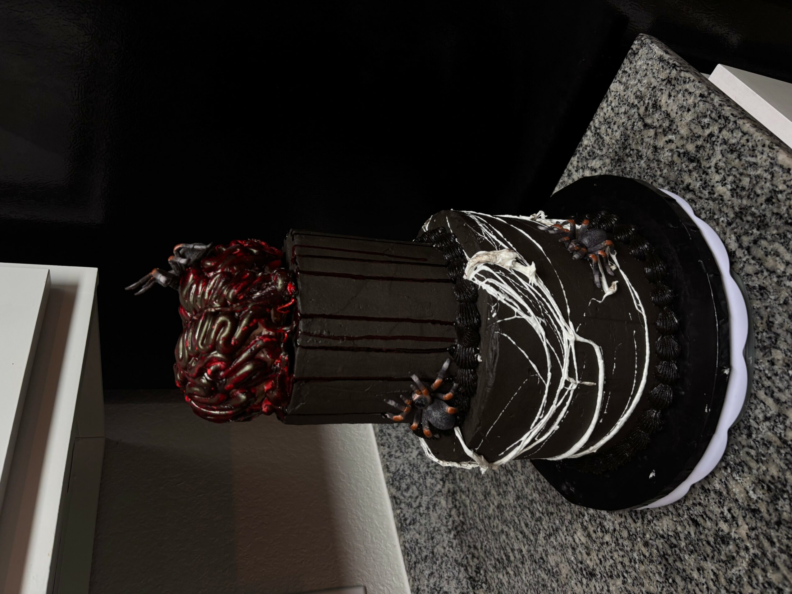 A multi-tiered black and chocolate cake with decorative elements, placed on a gray surface, with a dark background.