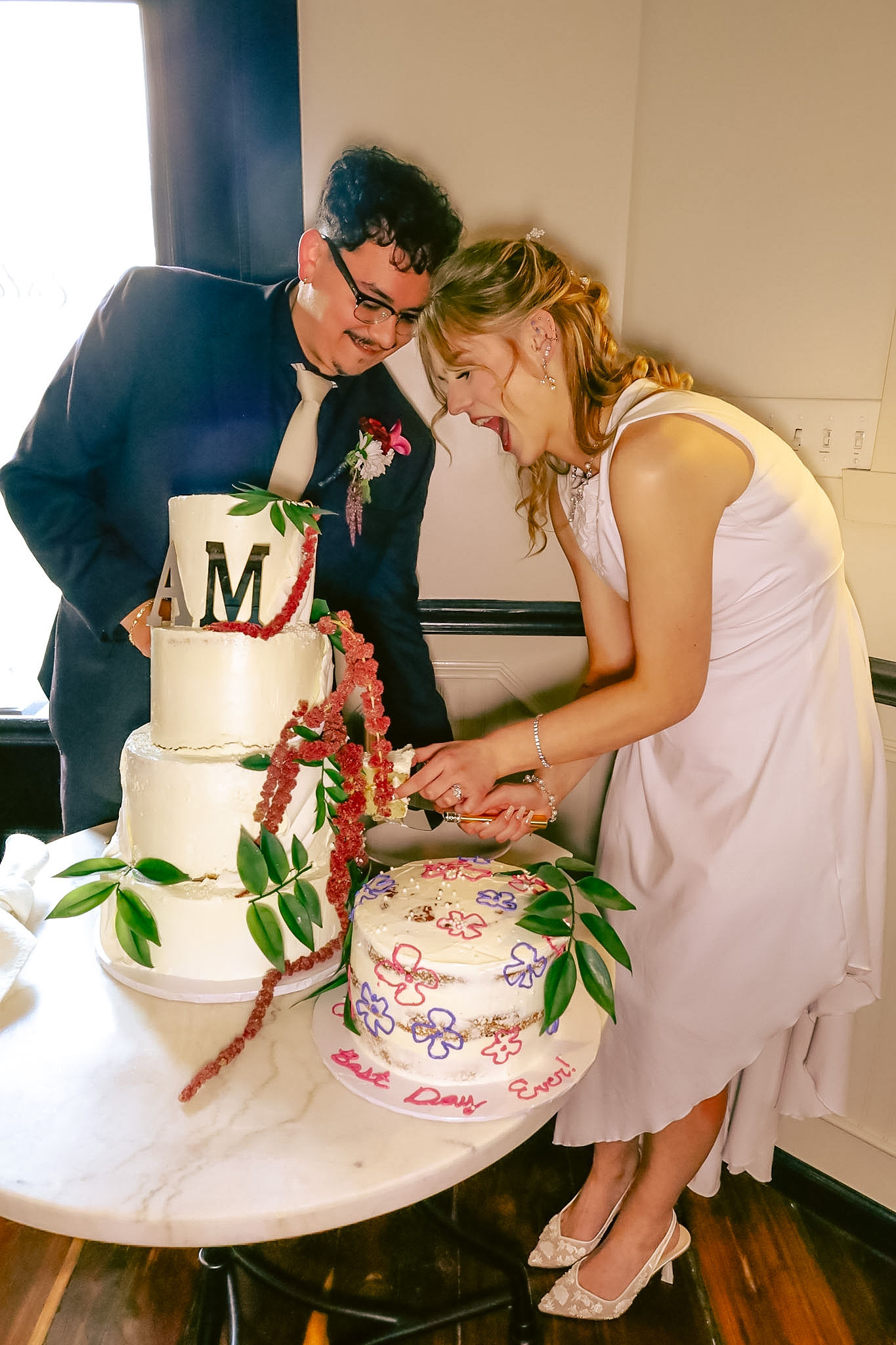 A couple in formal attire cutting a wedding cake together, with a three-tiered cake decorated with flowers and greenery on a table.