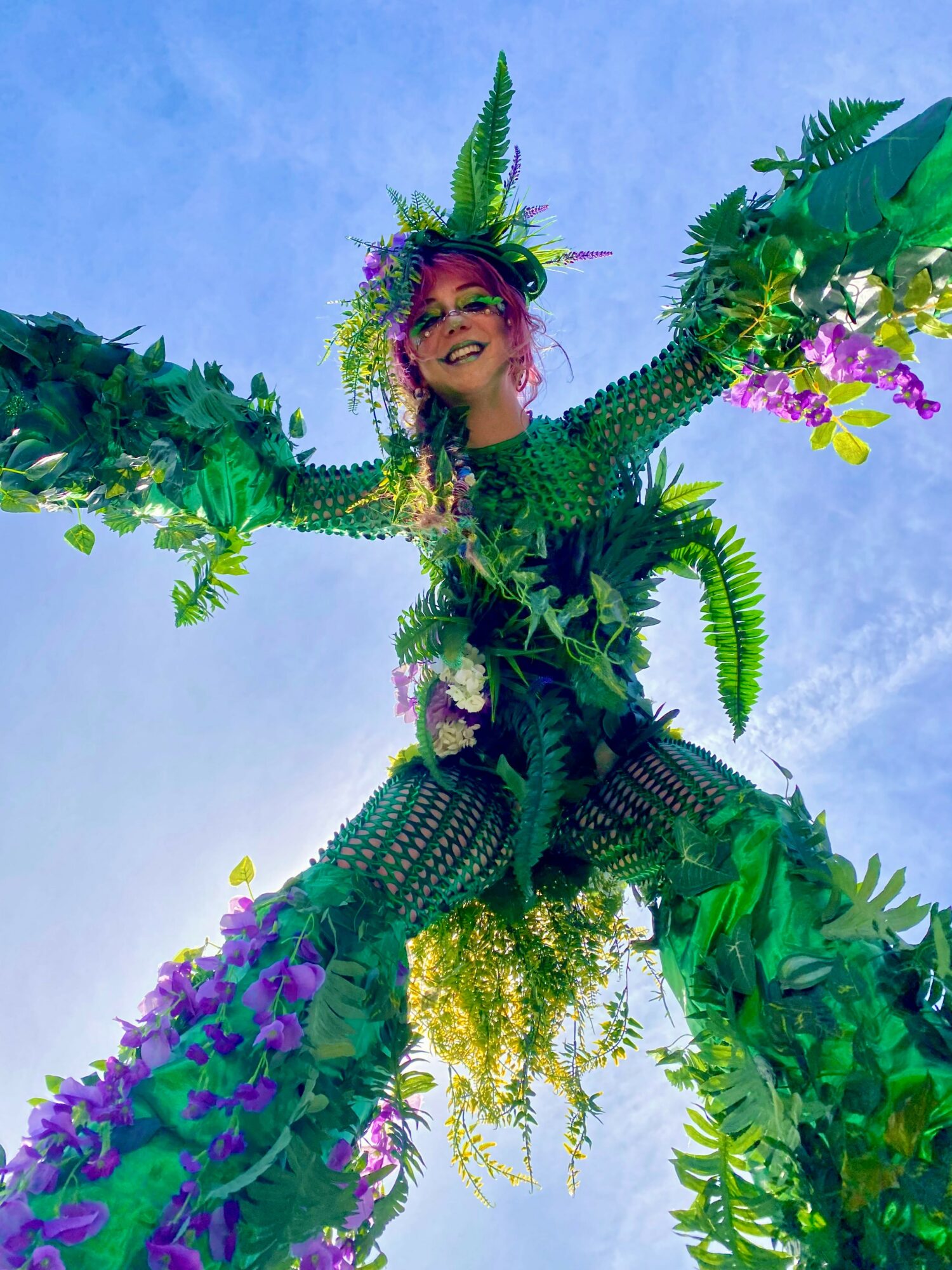 Person in elaborate green and purple costume with plant-like accessories, smiling against blue sky.