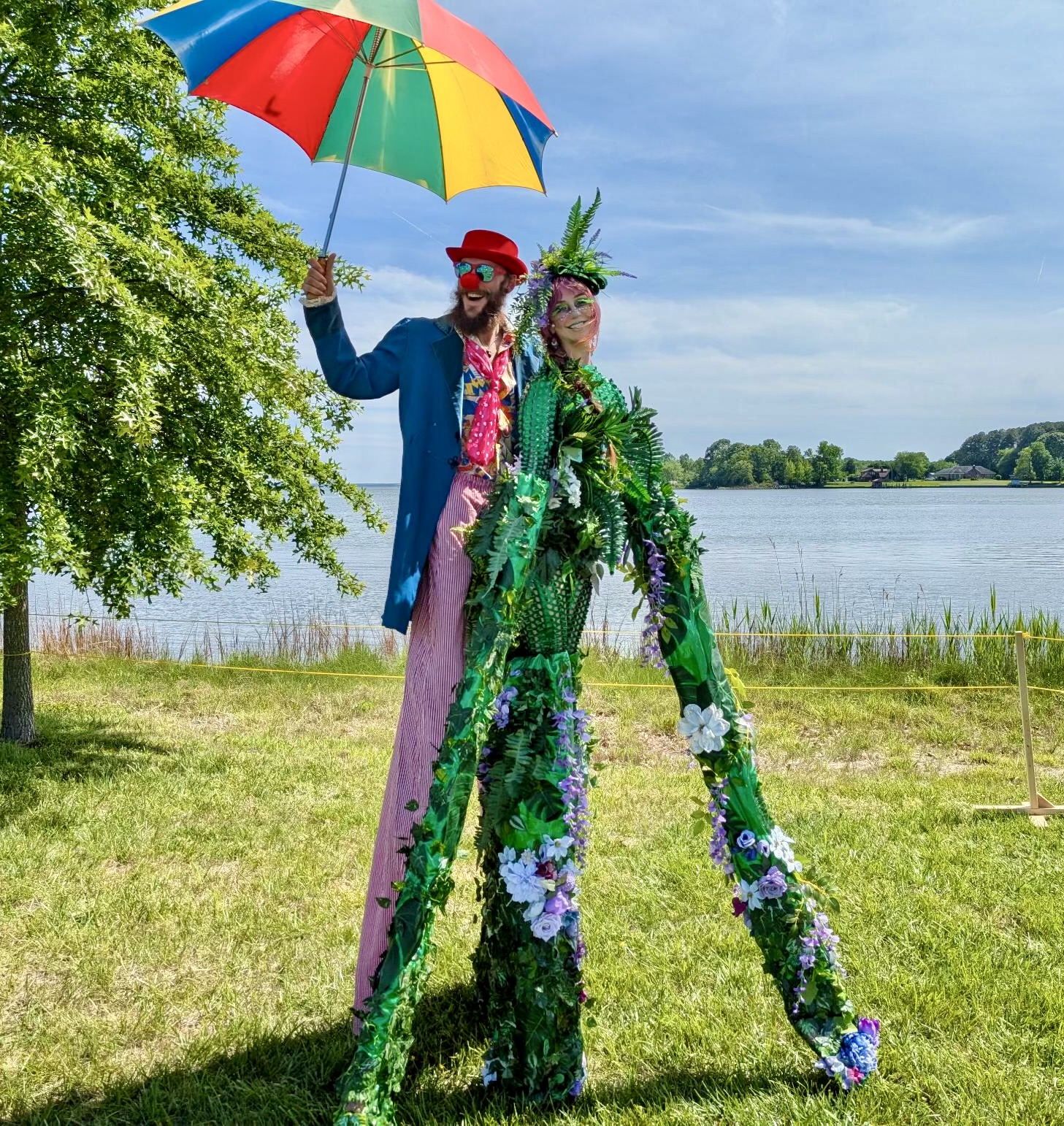 Two people dressed in colorful costumes stand outdoors near a lake, one holding a rainbow umbrella, with trees and water in background.