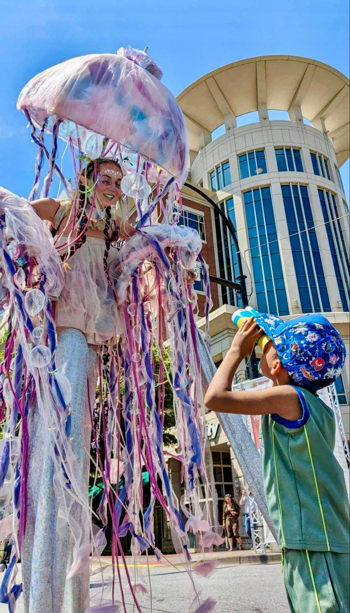 Child with a blue hat takes a photo of a person on a float with colorful ribbons and an umbrella, city buildings in background.