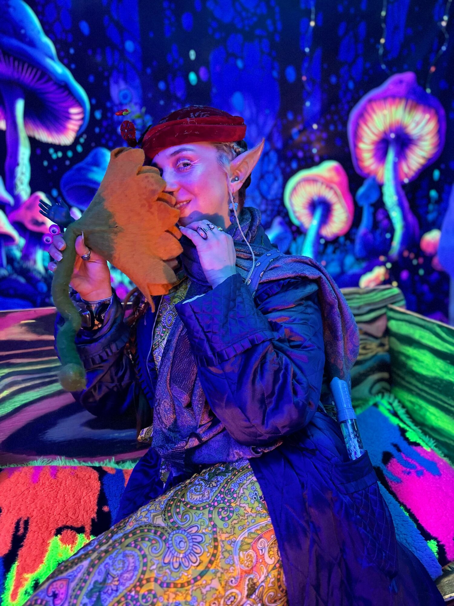 Person with pointed ears and red headband holding a plush toy, surrounded by colorful, glowing mushrooms in a fantasy setting.