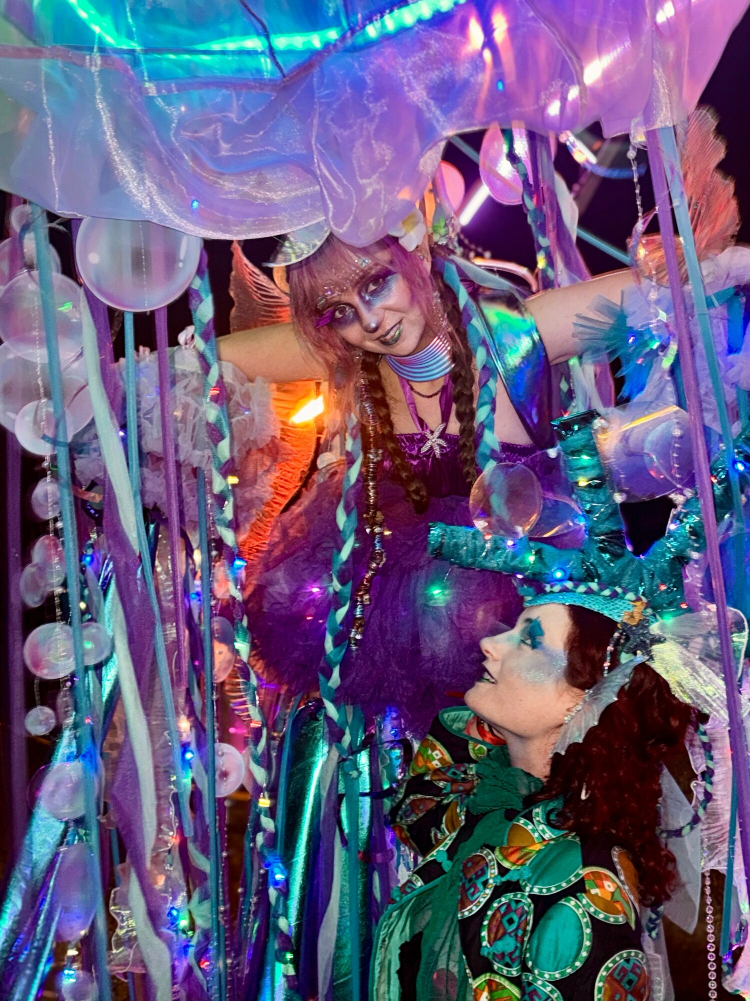 Two performers in colorful costumes with makeup and accessories, surrounded by lights and decorations, in a festive setting.