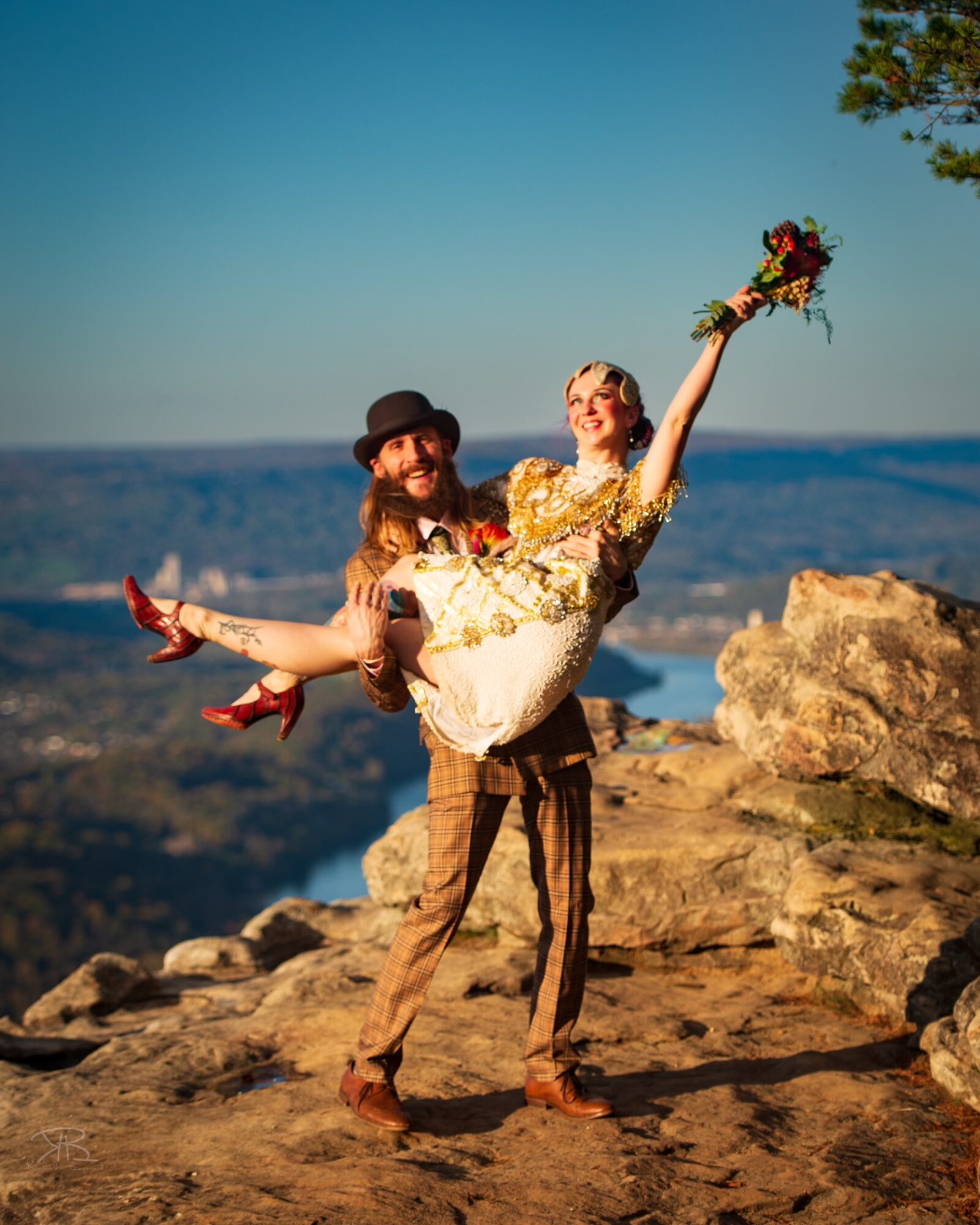 Man in brown pants and hat holding a woman in a wedding dress with a bouquet, outdoors on rocky terrain with a scenic background