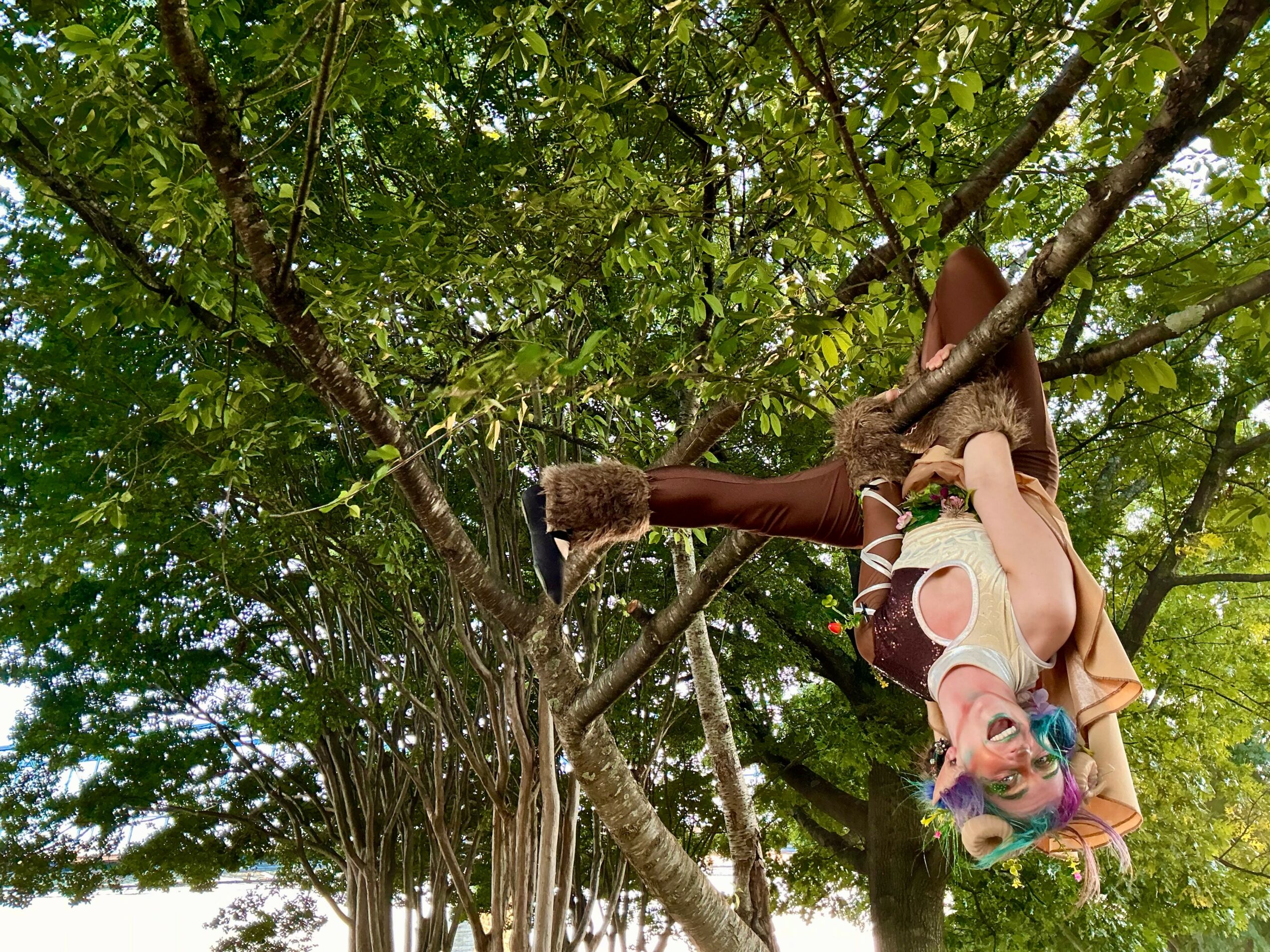 Person hanging upside down from a tree branch, surrounded by green leaves, wearing colorful clothing and accessories.