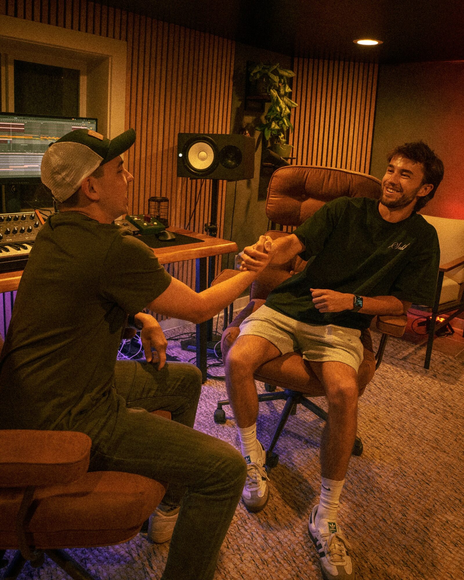 Two men in a recording studio, one sitting and the other standing, shaking hands and smiling, with audio equipment in background.