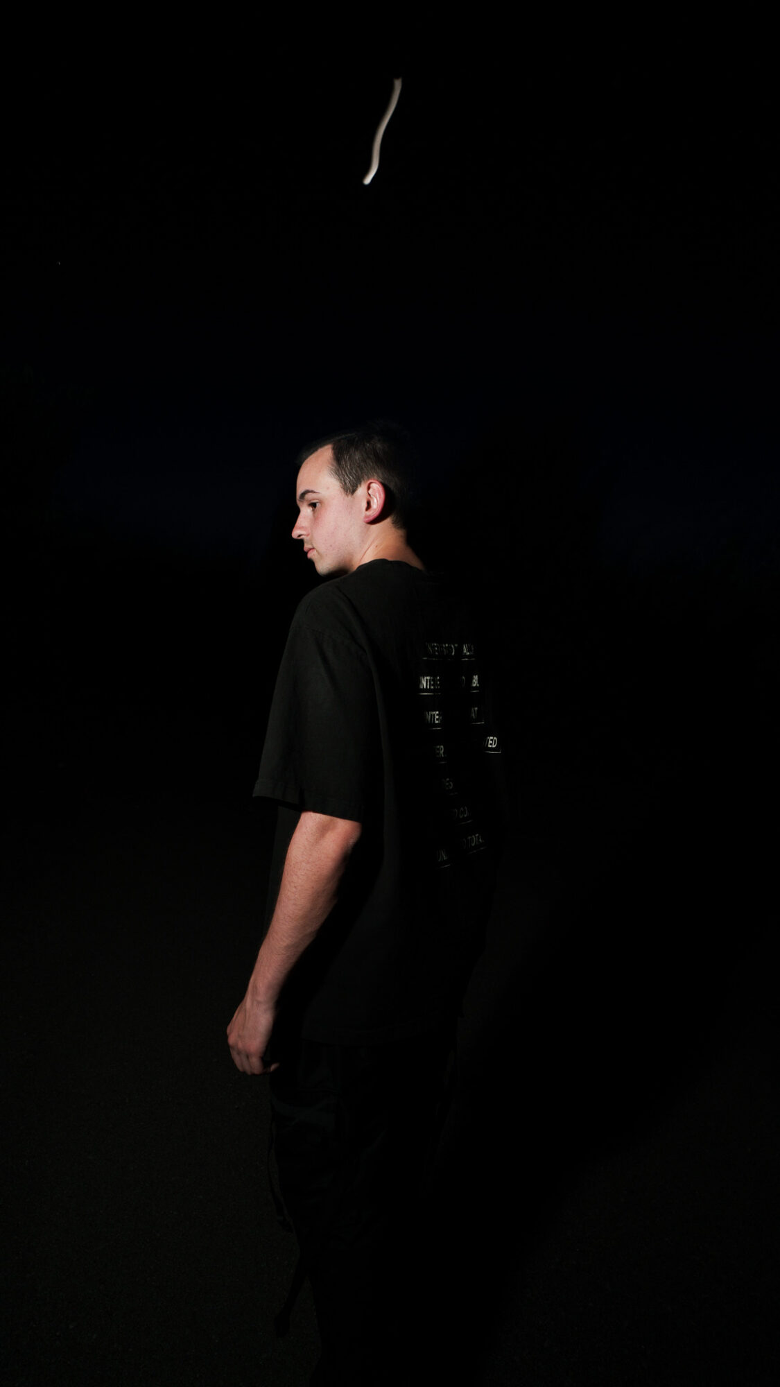 Side view of a person standing in a dark space, wearing a black shirt, with a dark background and a small light above.