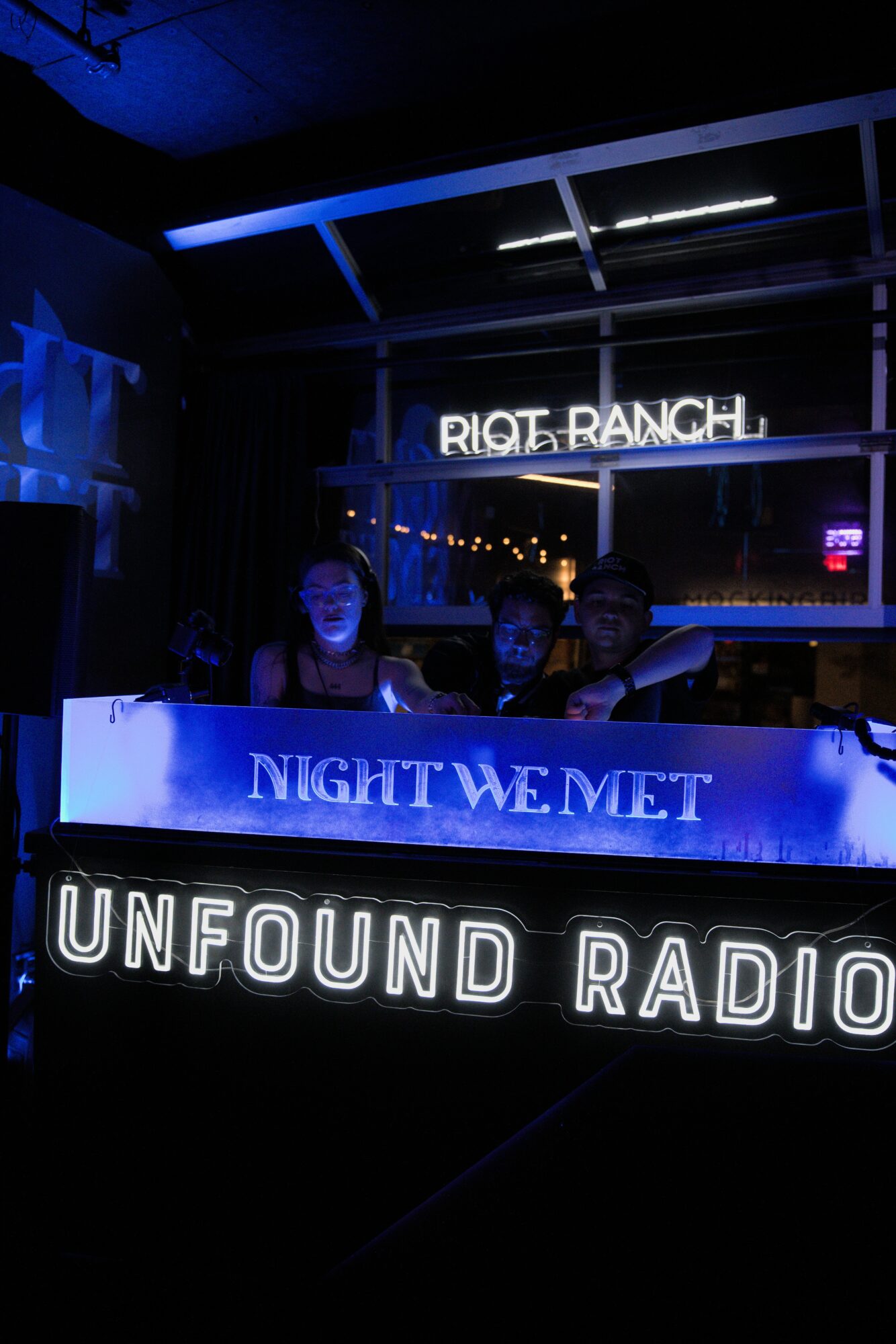 Neon signs reading 'RIOT RANCH,' 'NIGHT WE MET,' and 'UNFOUND RADIO' in a dark indoor setting with three people in the background.