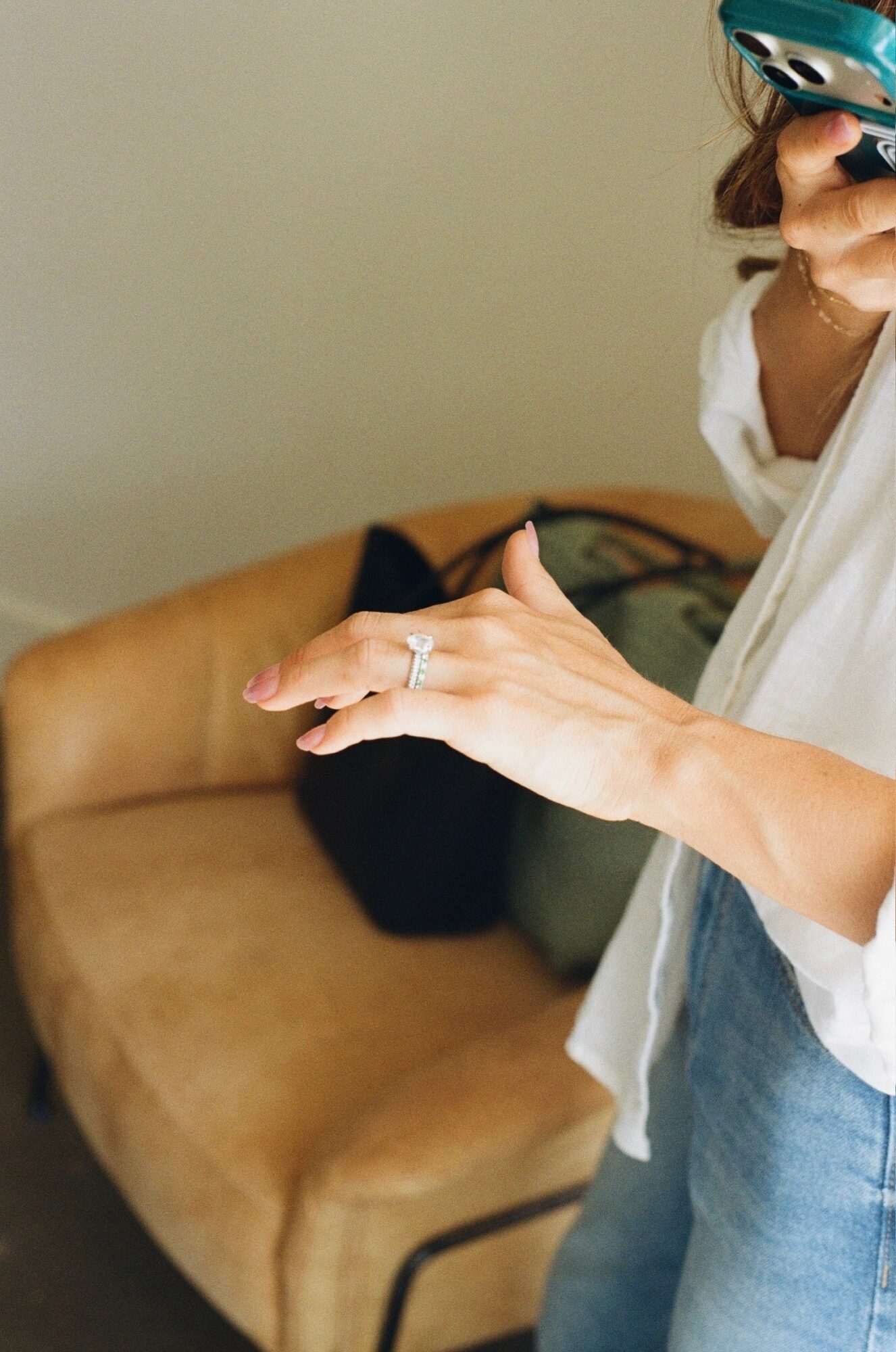 Person holding a phone with a ring on their finger, standing near a beige sofa, wearing a white shirt and jeans.