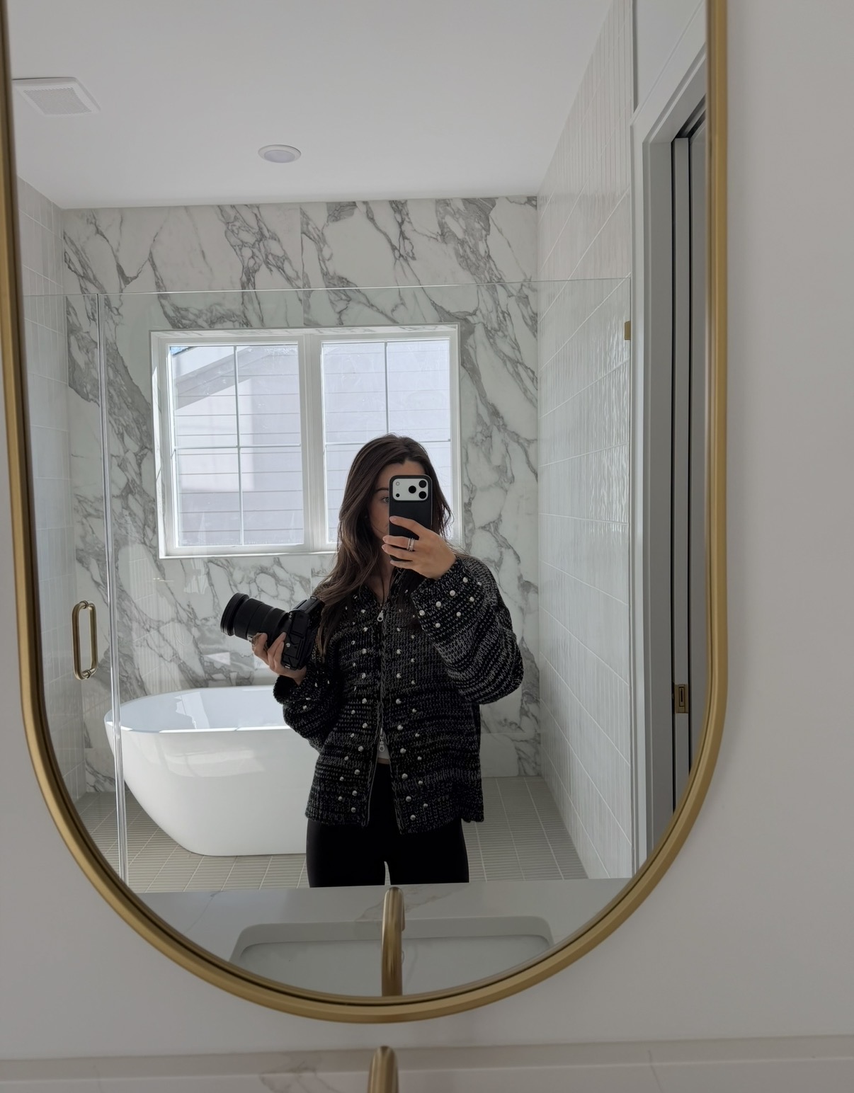 Person taking a mirror selfie in a bathroom with marble walls and a window, holding a phone and a small object.