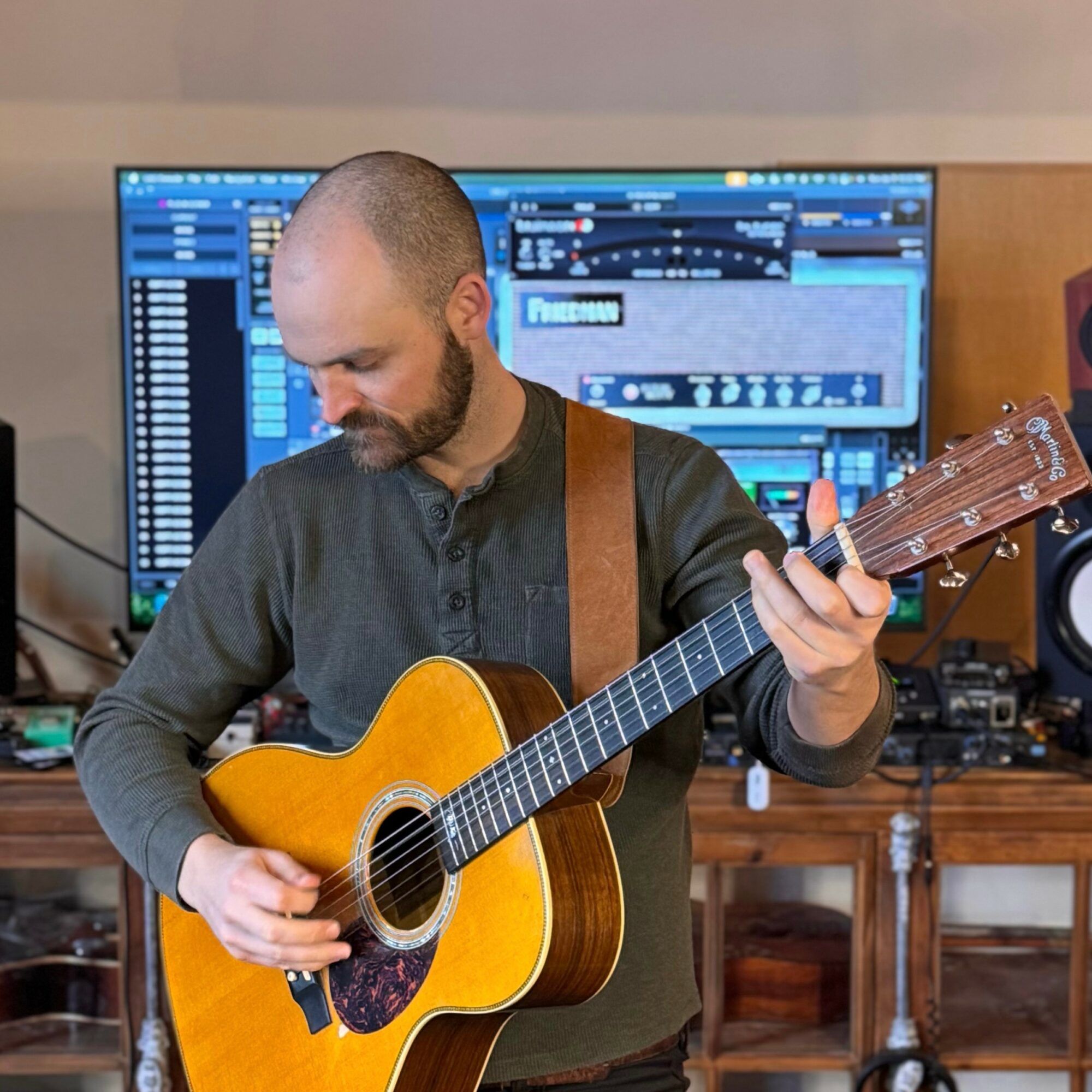 Man playing an acoustic guitar in a recording studio with computer screens in the background.