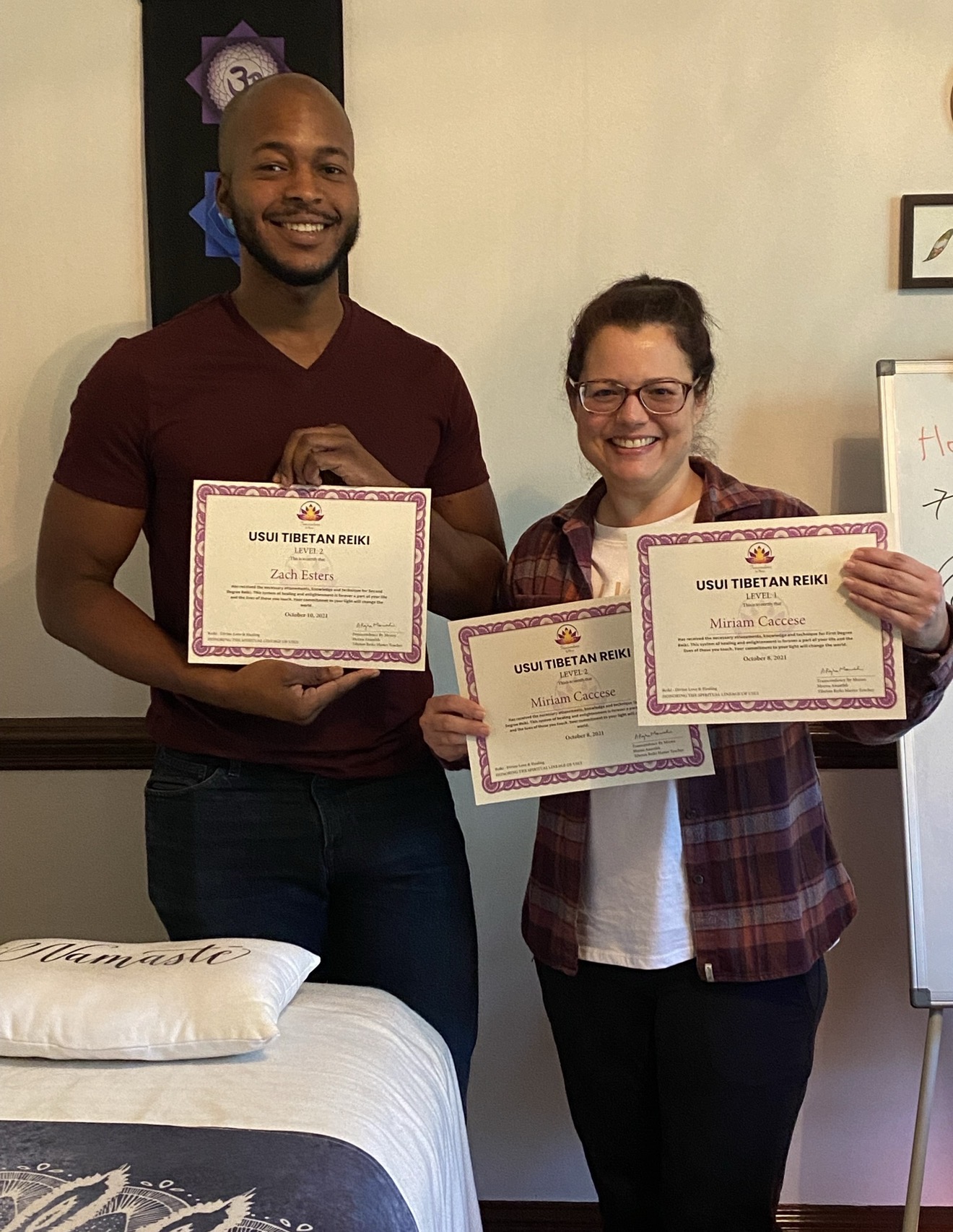 Two people holding certificates, smiling indoors, with a bed and whiteboard in the background.
