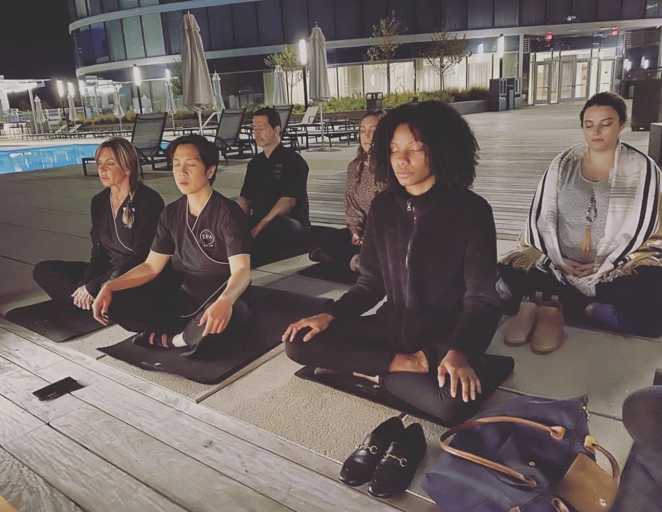 Group of six people practicing meditation outdoors at night, sitting cross-legged on mats near a pool, with a building in the background.