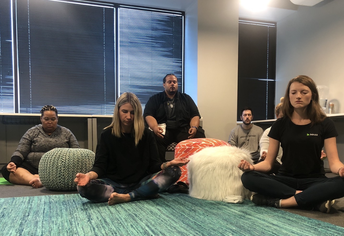 Group of five people practicing meditation or yoga in a room with large windows, sitting cross-legged on a green rug.