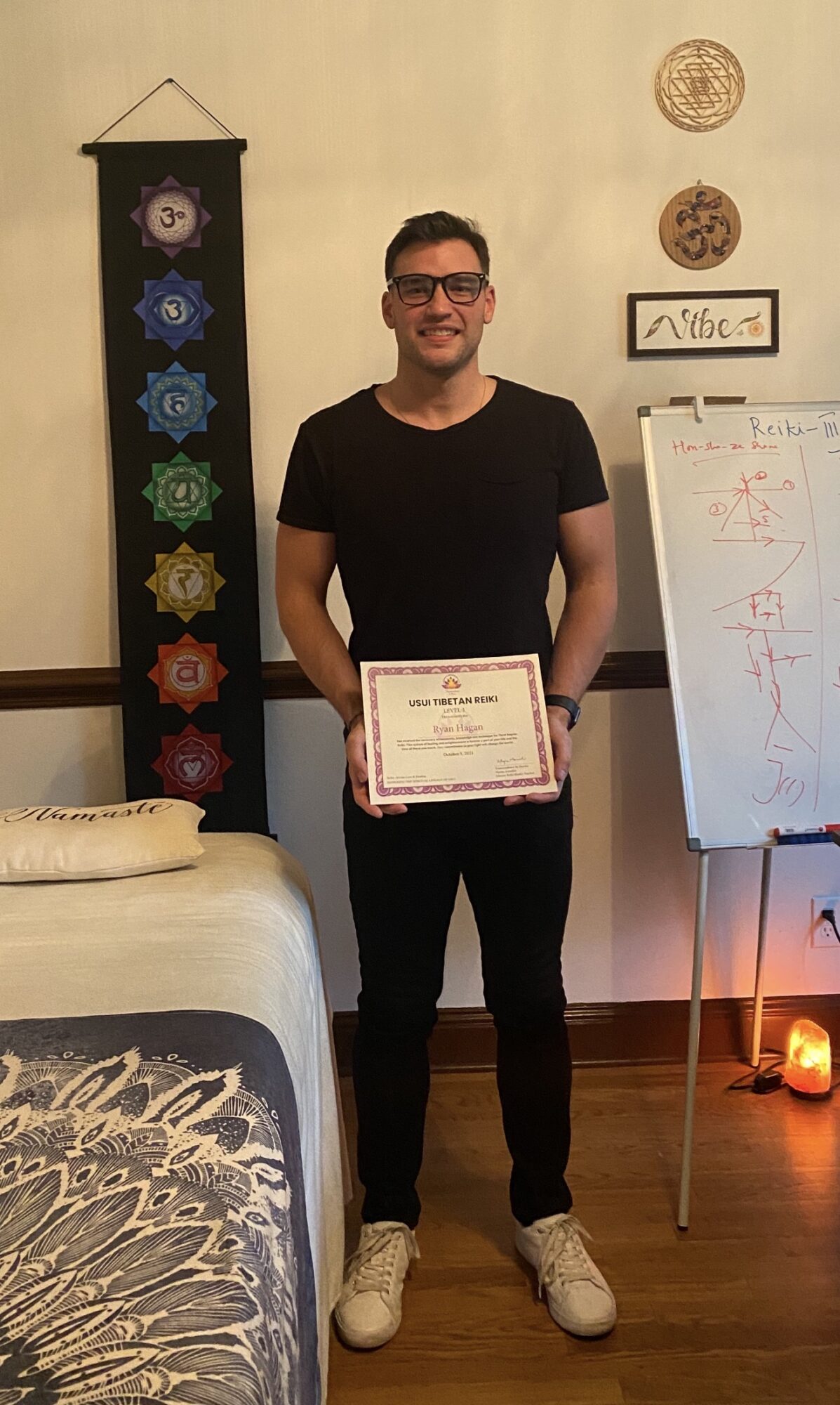 Young man holding a certificate, standing in a room with wall decorations and a whiteboard, smiling.
