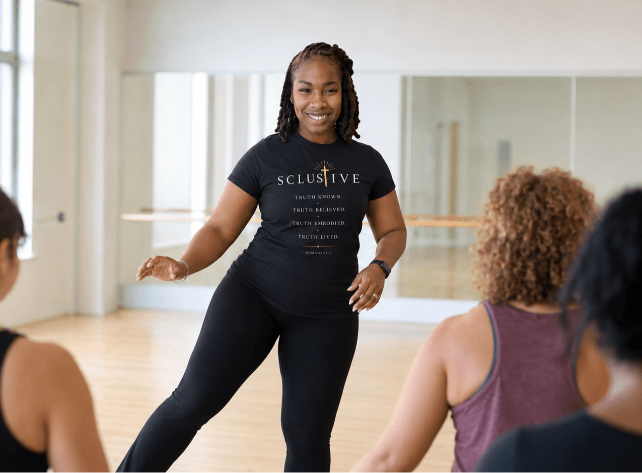 Woman leading a fitness class, smiling, in a studio with mirrors and wooden floor, with three participants visible from behind.