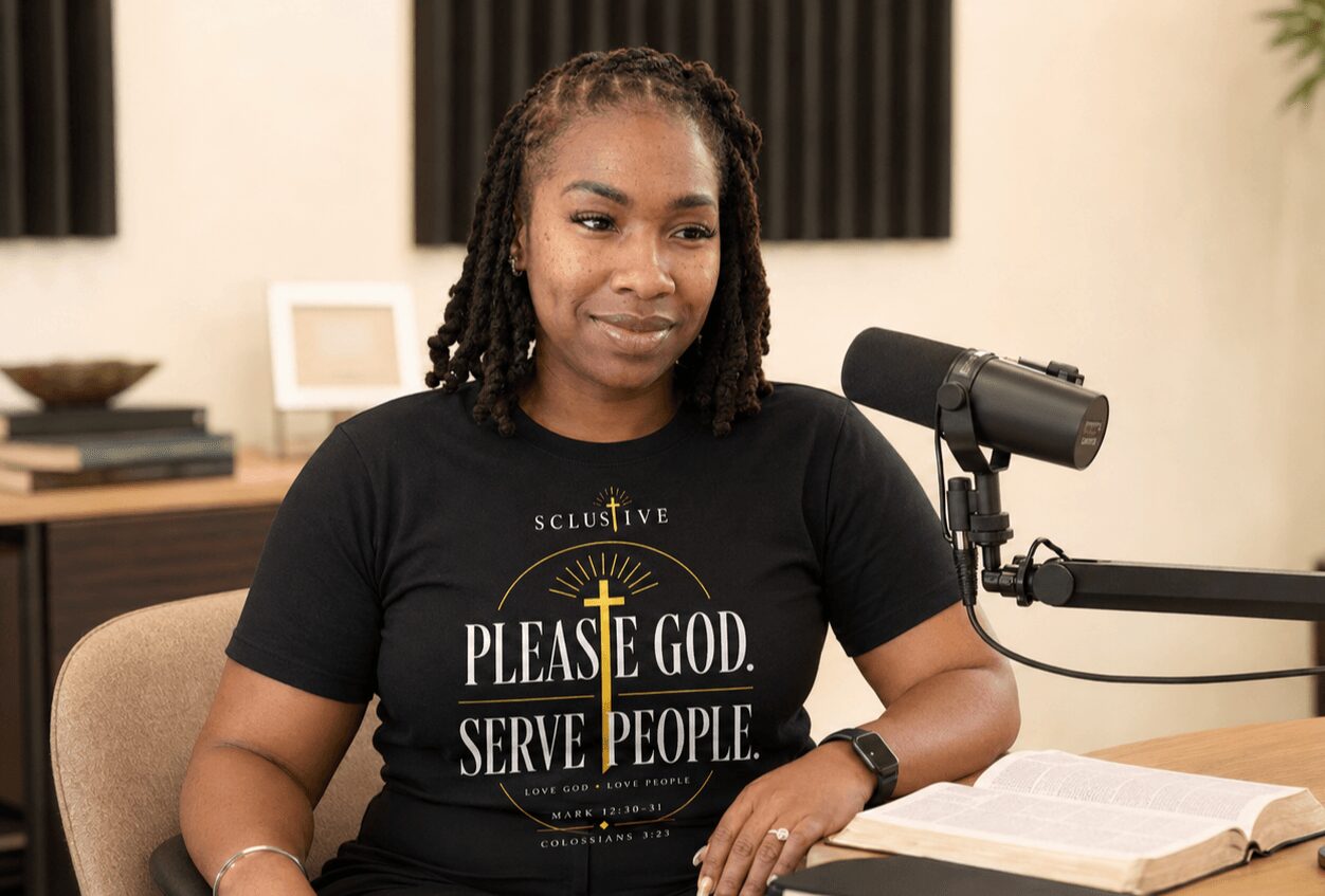 Woman with dreadlocks sitting at a desk with a microphone and open book, in a recording studio or podcast setting.