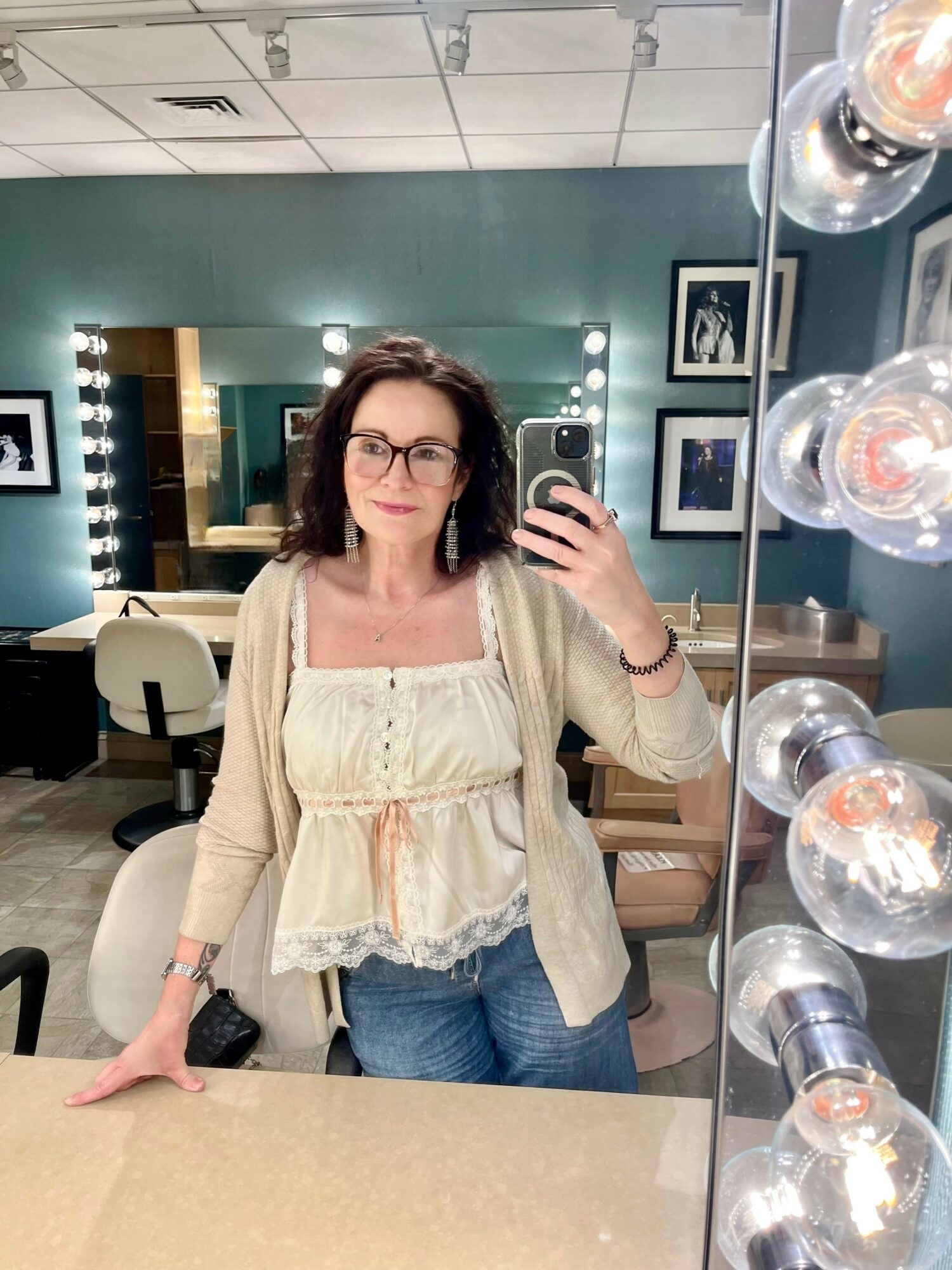 Woman with glasses taking a mirror selfie in a dressing room, wearing a beige cardigan and white top, with vanity lights visible.