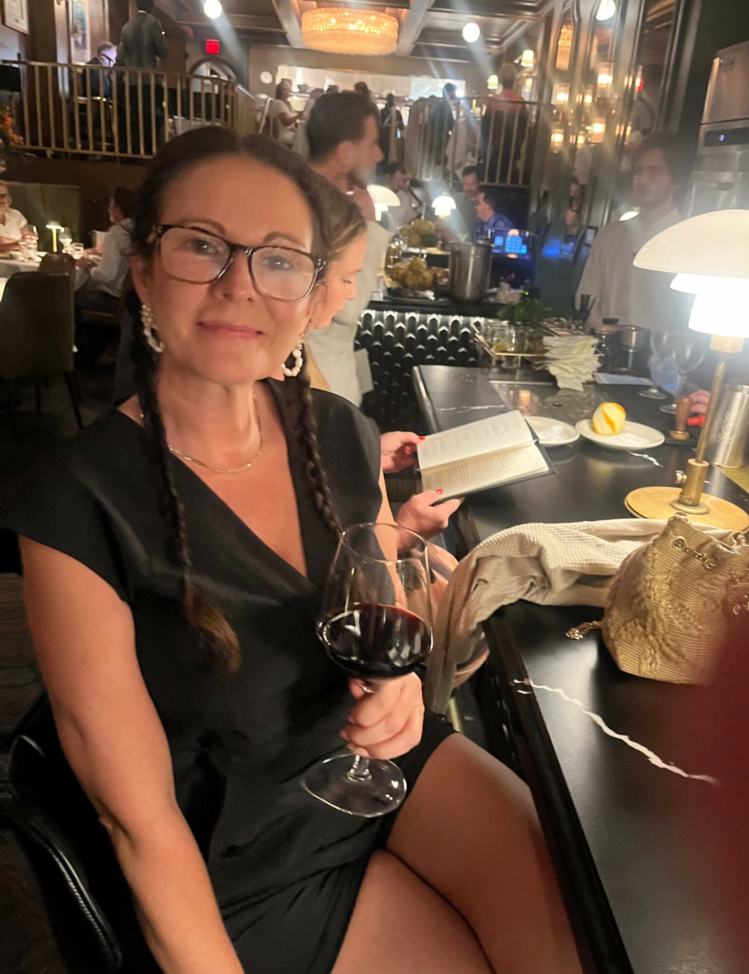 Woman with glasses holding a glass of red wine in a restaurant, with a book and a lamp on the table.