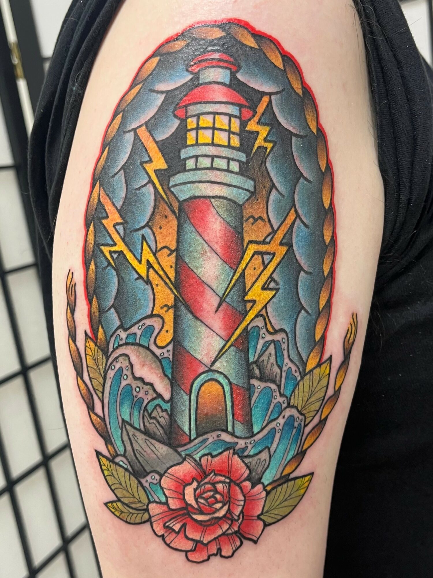 Lighthouse with red and white stripes, surrounded by stormy clouds, lightning, and a pink rose at the base, on upper arm.