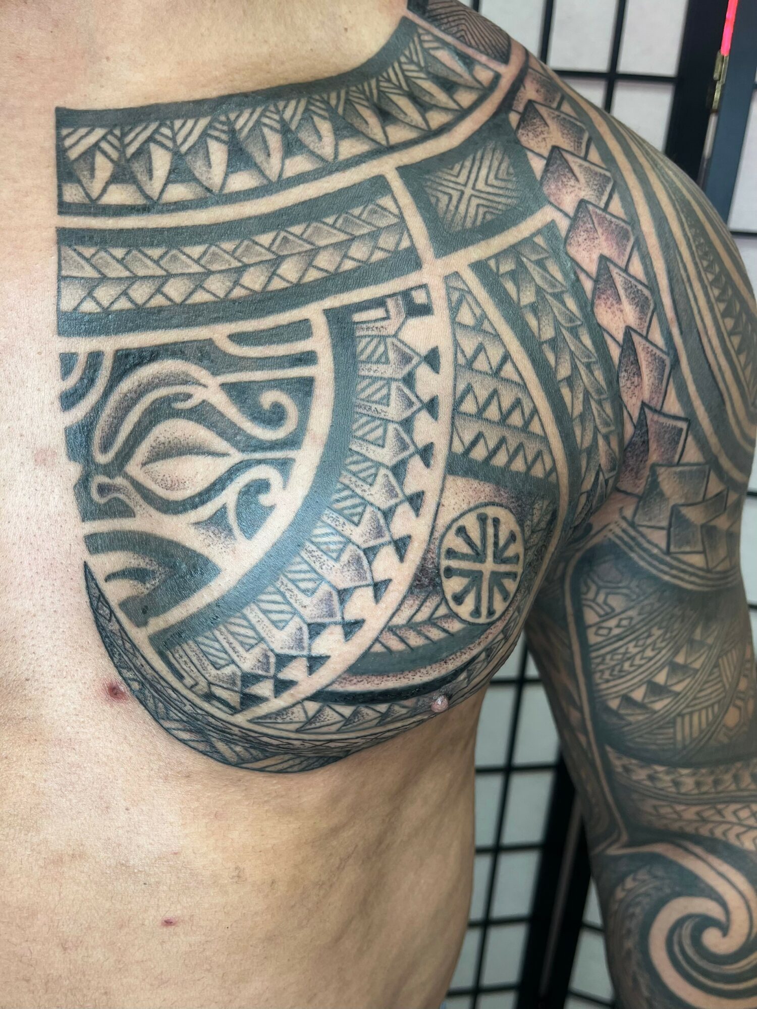 Close-up of a person's shoulder and upper arm with intricate black and gray Polynesian-style tattoo patterns.