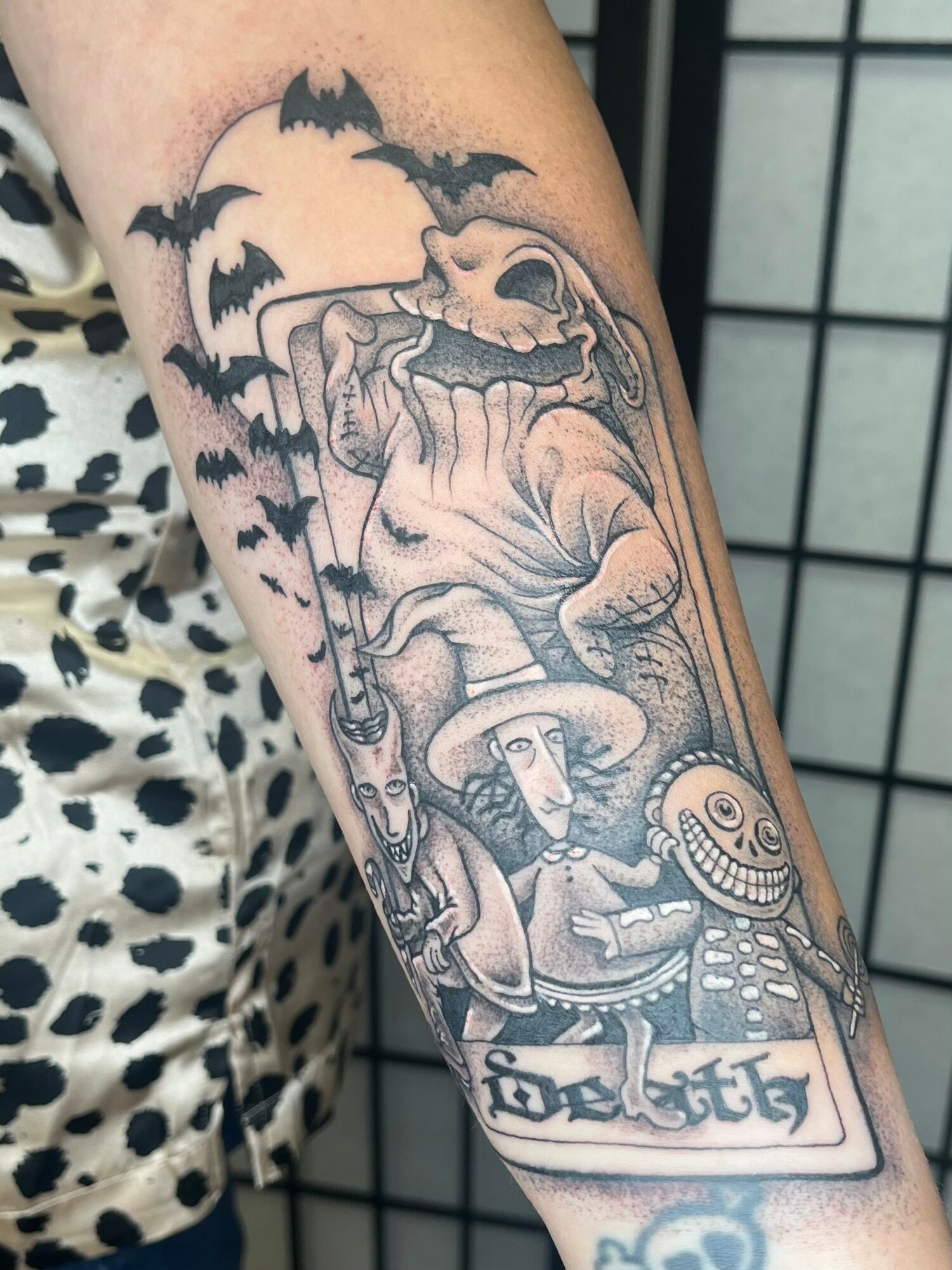 Tattoo of a skeleton, a scarecrow, and bats flying, with a banner reading 'Dios' and a grinning skull, in black and gray.