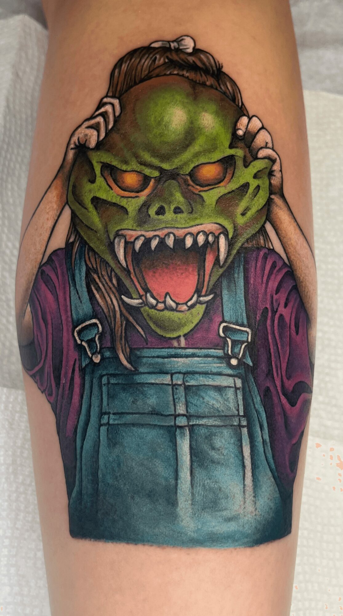 Tattoo of a green monster with orange eyes, sharp teeth, wearing overalls and a purple shirt.