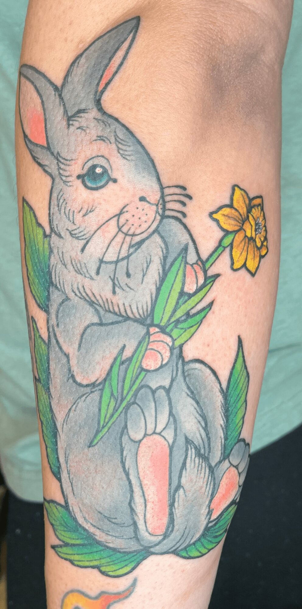 Colorful tattoo of a bunny holding a yellow flower, sitting among green leaves, on a person's arm.
