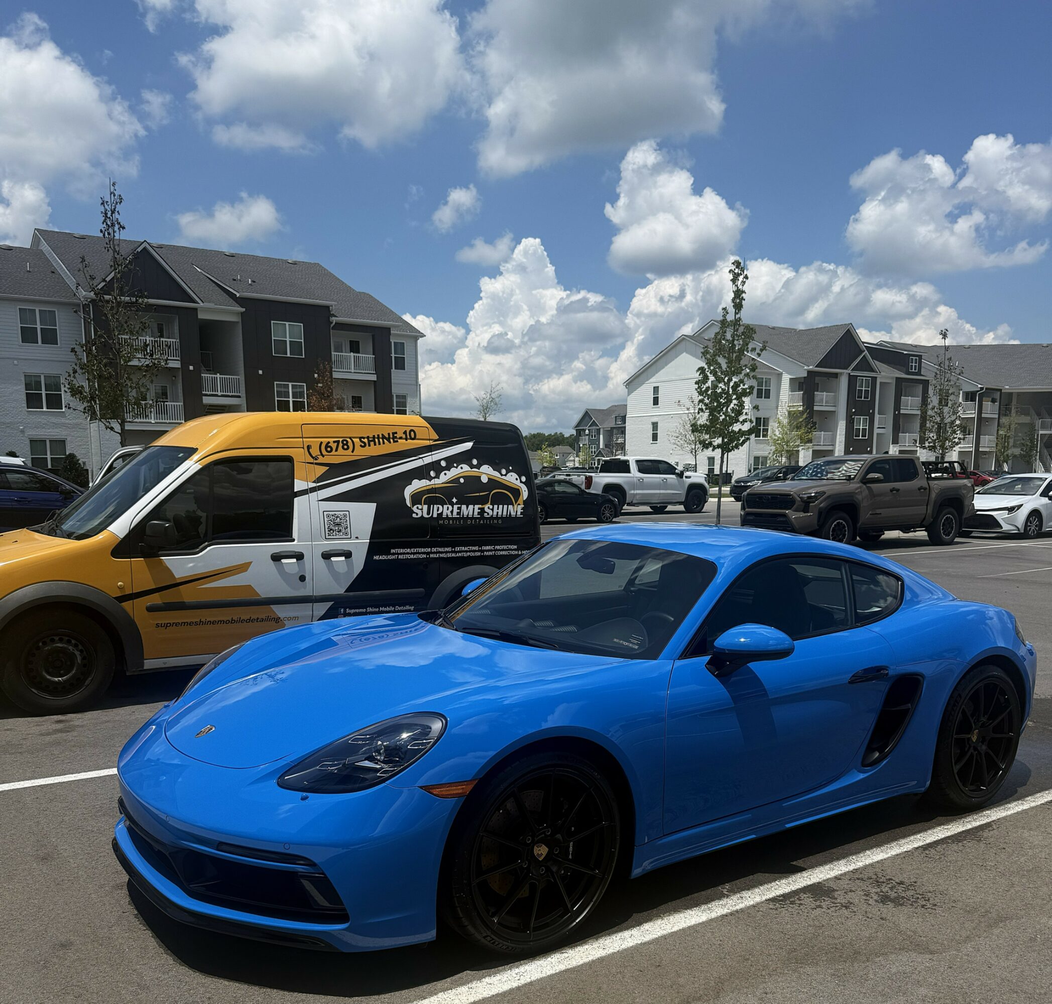 Blue sports car parked in a lot with apartment buildings and a yellow vehicle nearby under a partly cloudy sky.