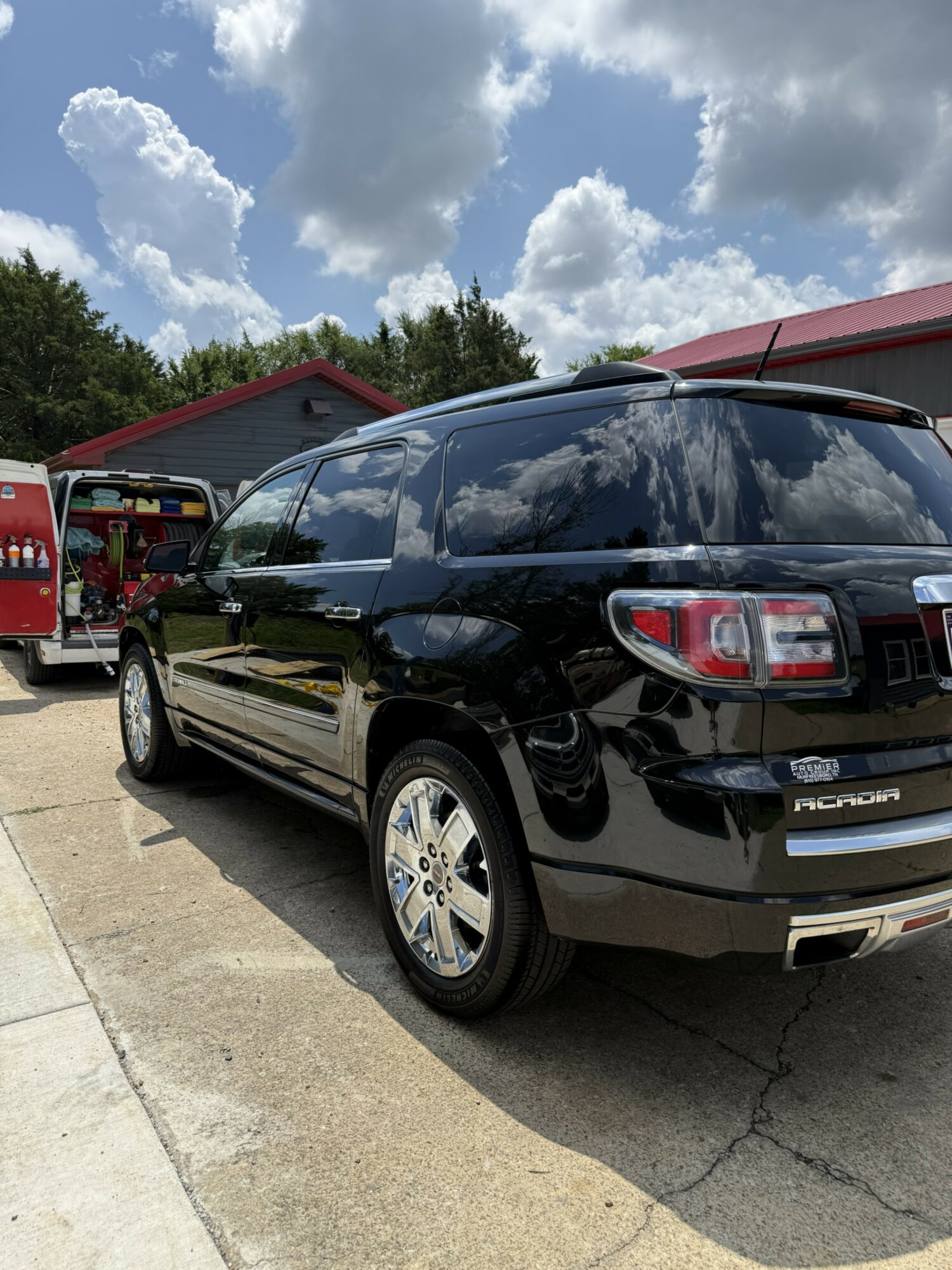 Black SUV parked outdoors with a red building and trees in the background, under a partly cloudy sky.