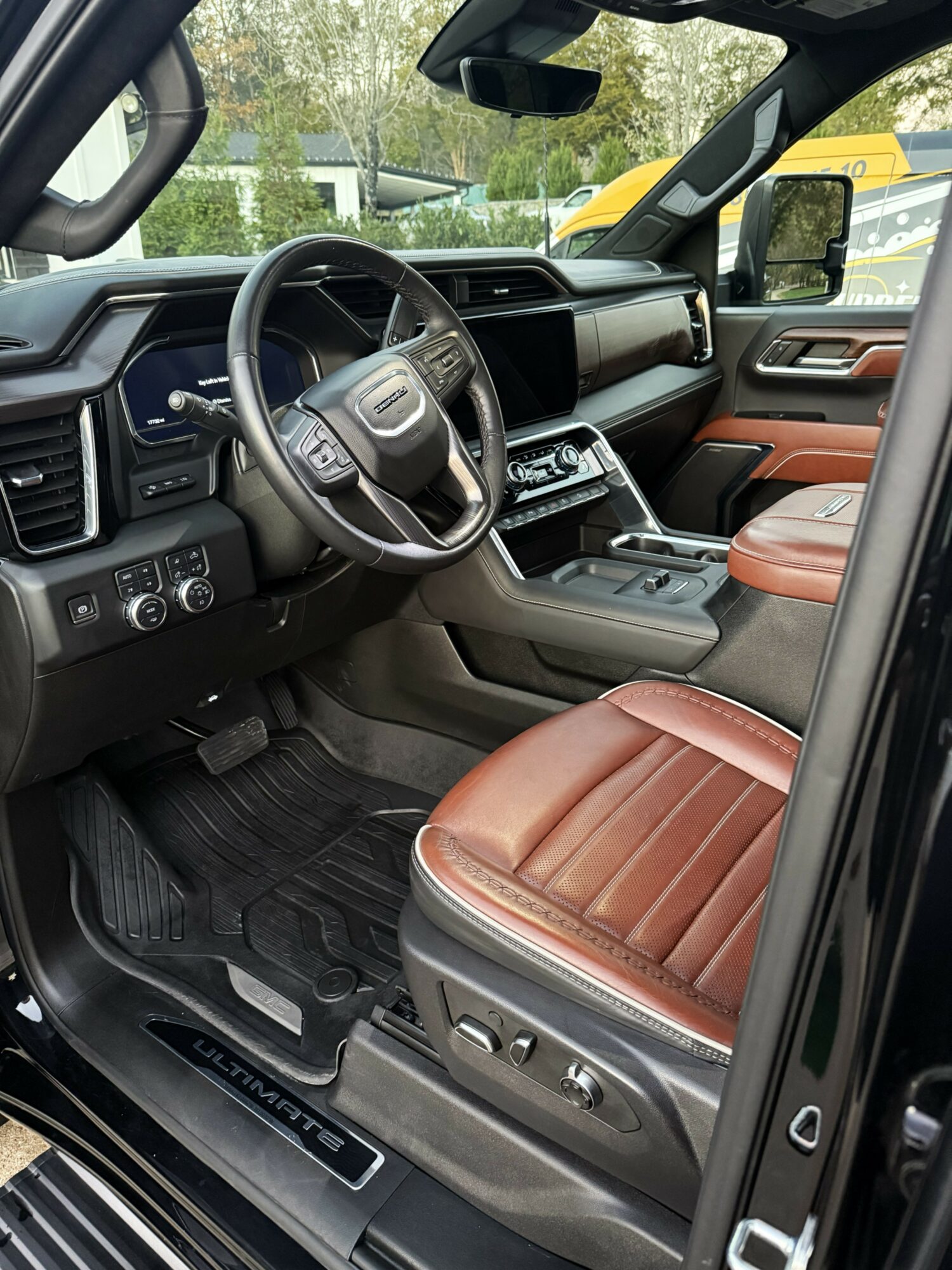 Interior of a vehicle showing a steering wheel, dashboard, and brown leather seat, with a view through the windshield of trees and a yellow vehicle outside.