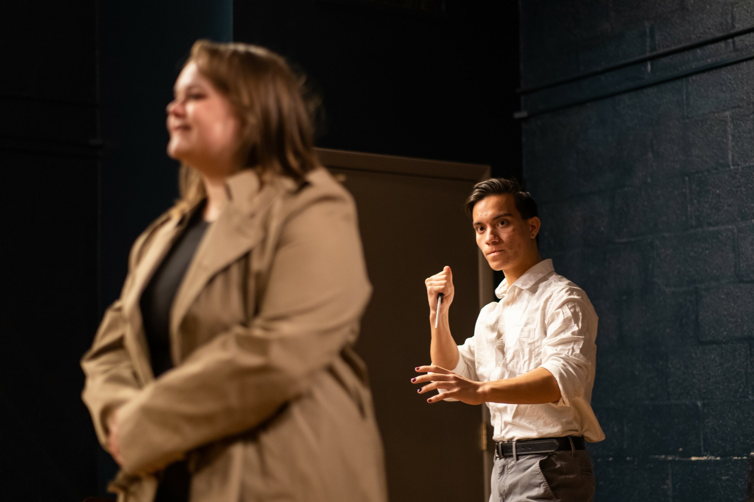 Two people on stage, one in the foreground with blurred face, the other in the background gesturing with hand, dark background.