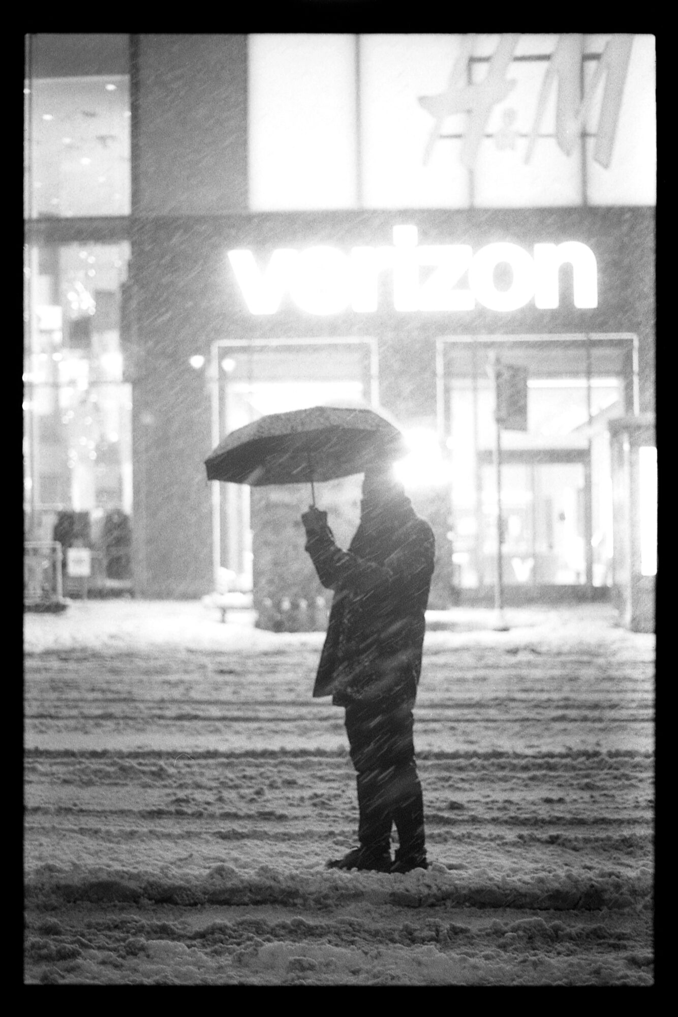 Person standing with umbrella on snowy street at night, Verizon store in background, black and white image.