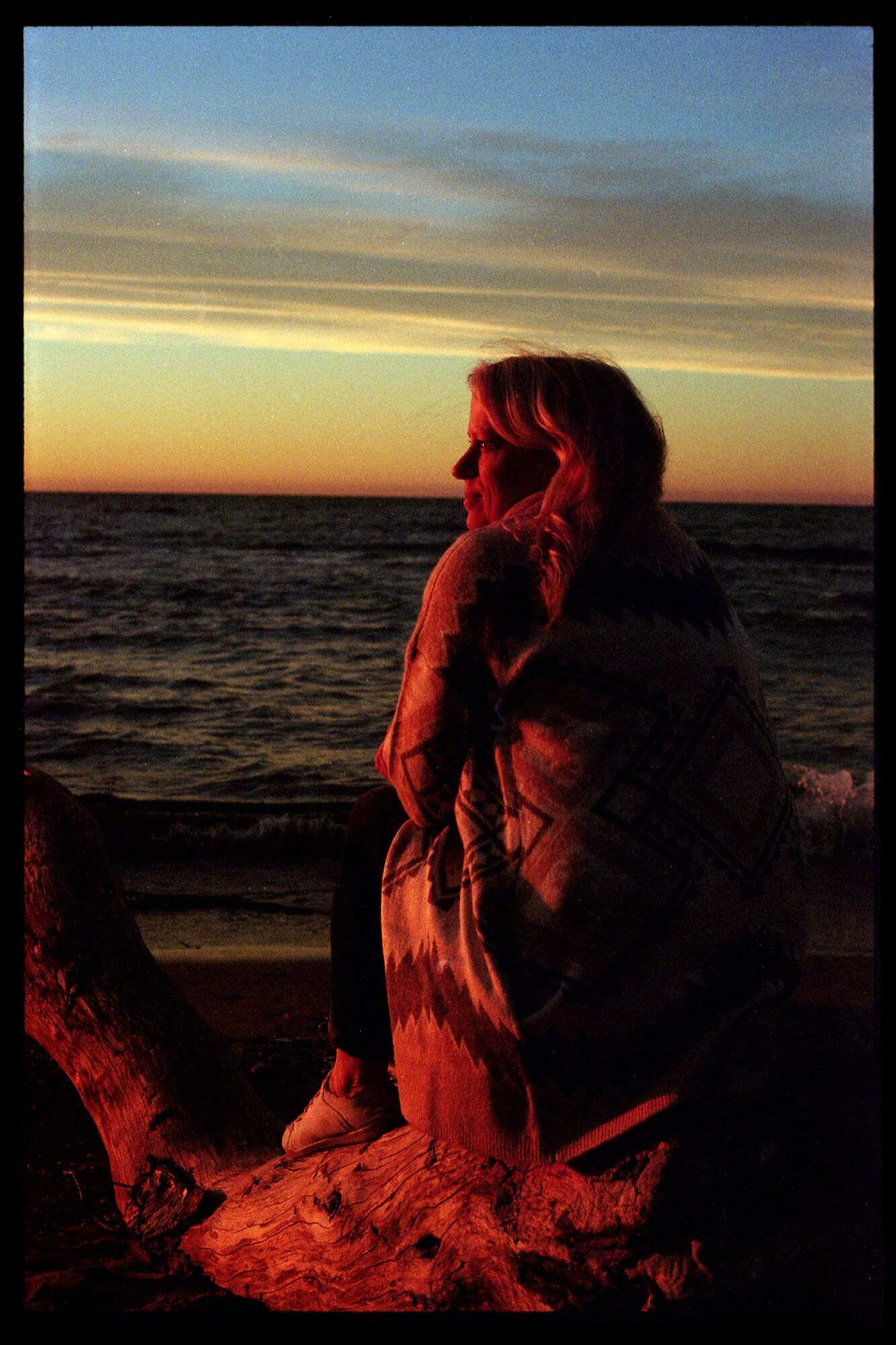 Person sitting on a beach at sunset, looking at the ocean, with a colorful sky and clouds.