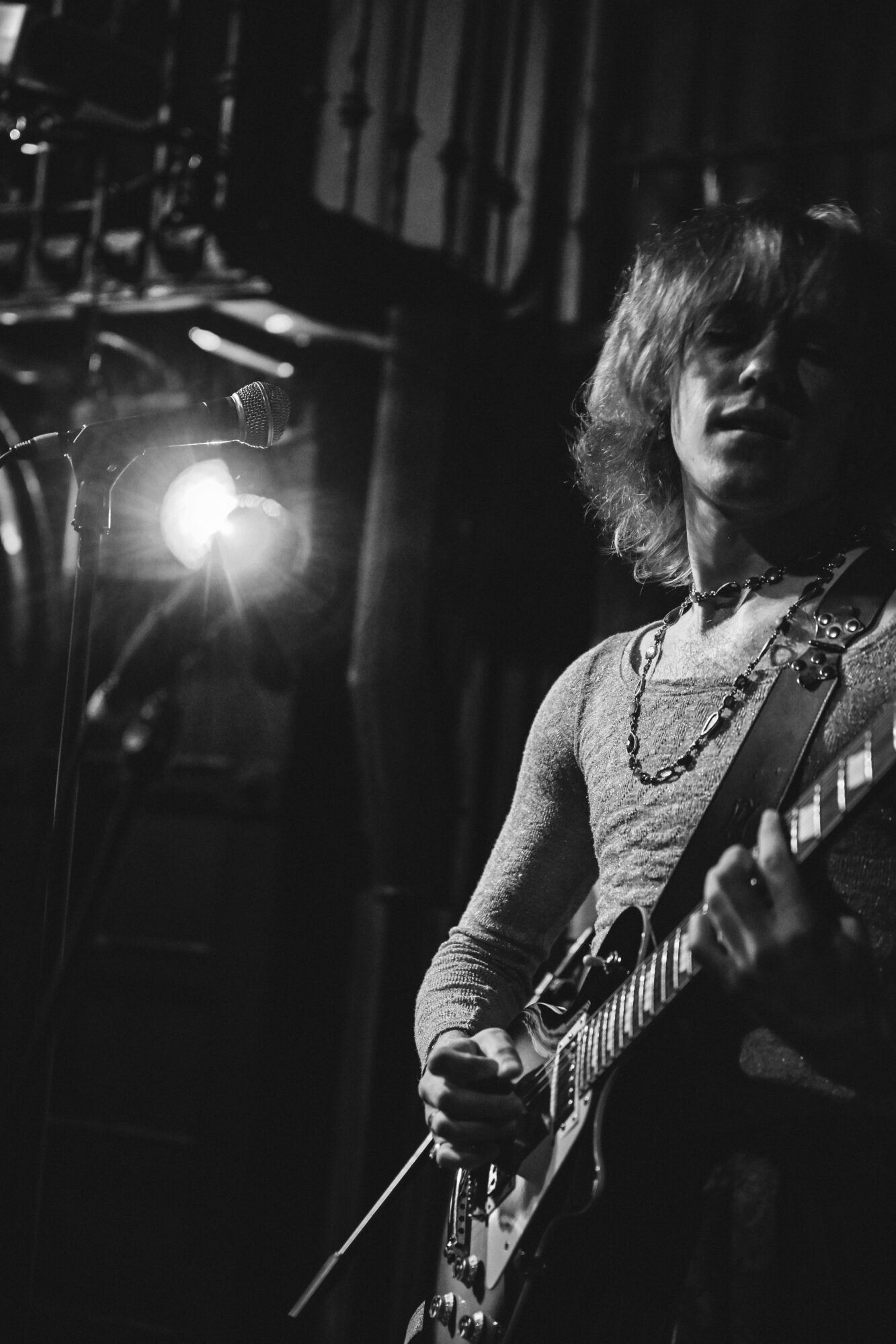 Person playing guitar on stage with a bright light behind them, dark background, shoulder-length hair, wearing a necklace.