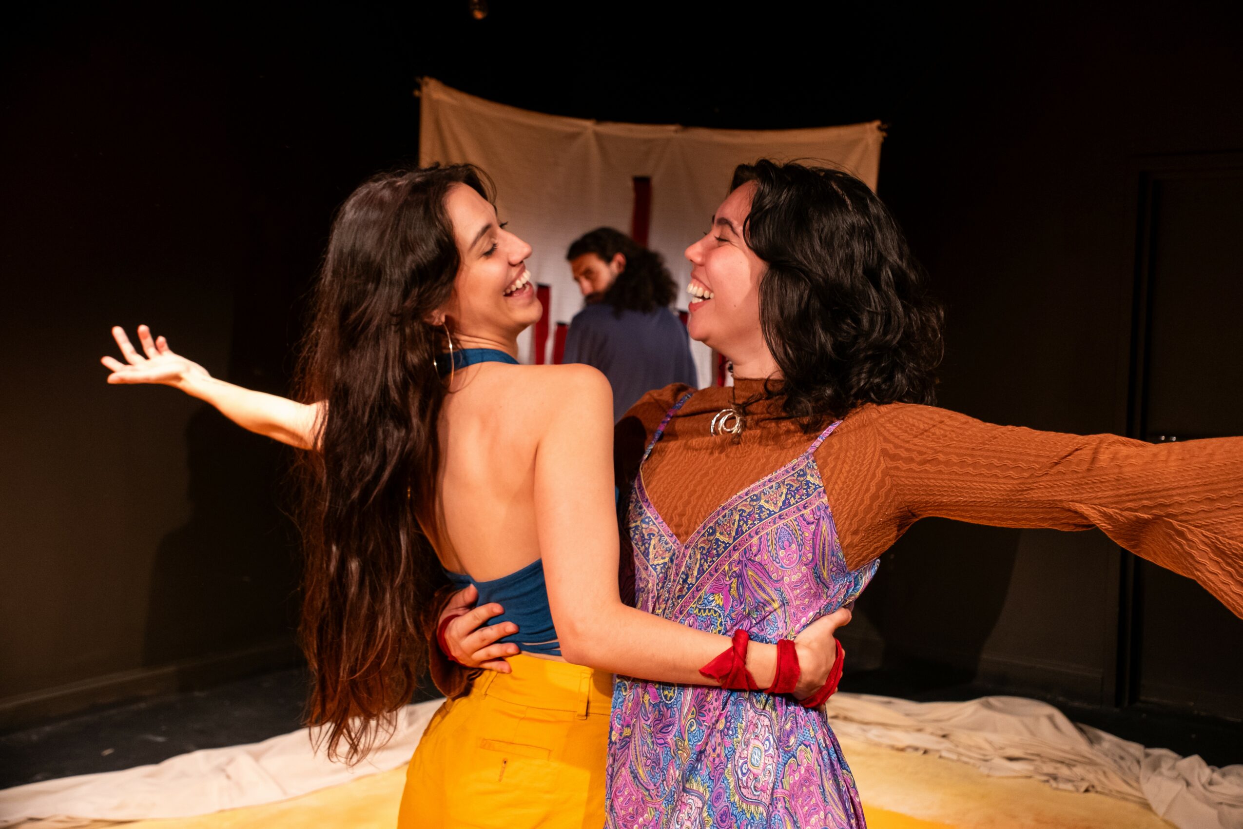 Two women smiling and embracing on stage, with a backdrop and other people in the background.
