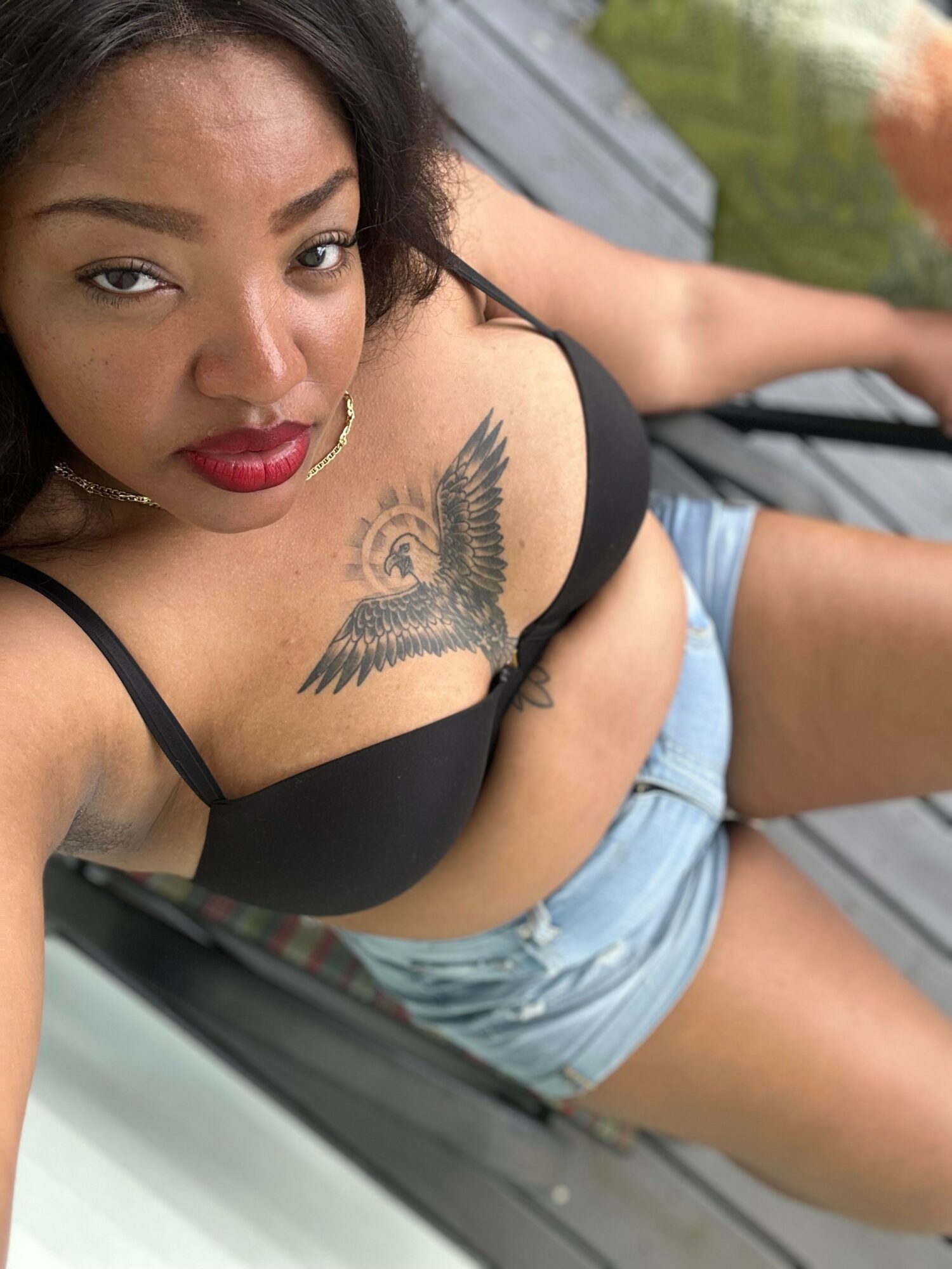 Woman with dark hair and red lipstick, wearing a black top, sitting outdoors with a tattoo of a bird on her chest.