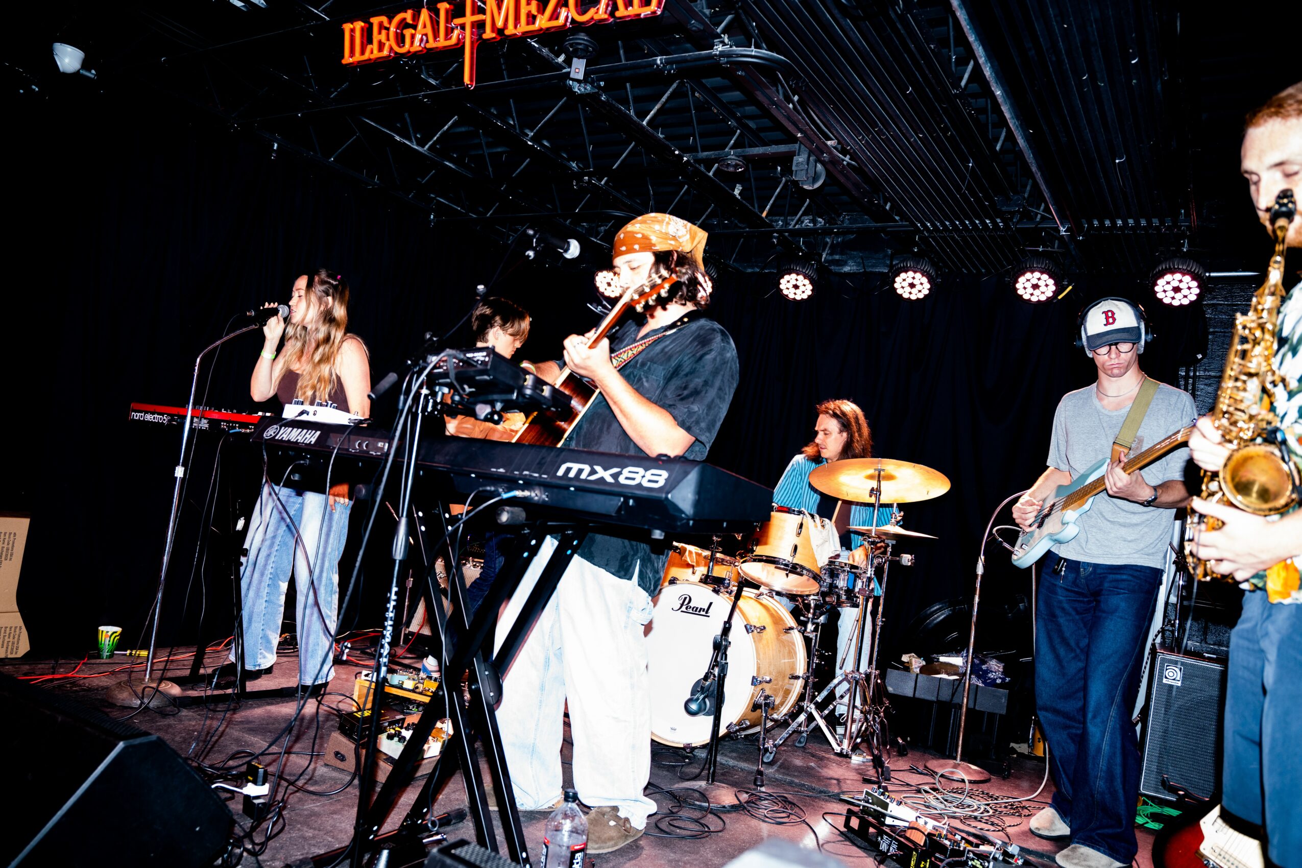 Band performs on stage with instruments, including keyboard, drums, guitar, and saxophone, under stage lights and a black backdrop.