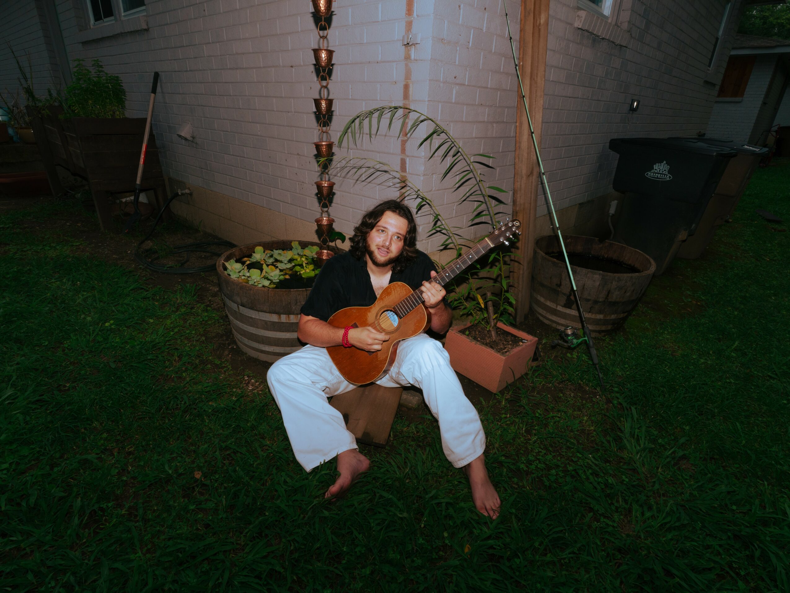 Person sitting outdoors playing guitar, surrounded by plants and garden items, with a house in the background.