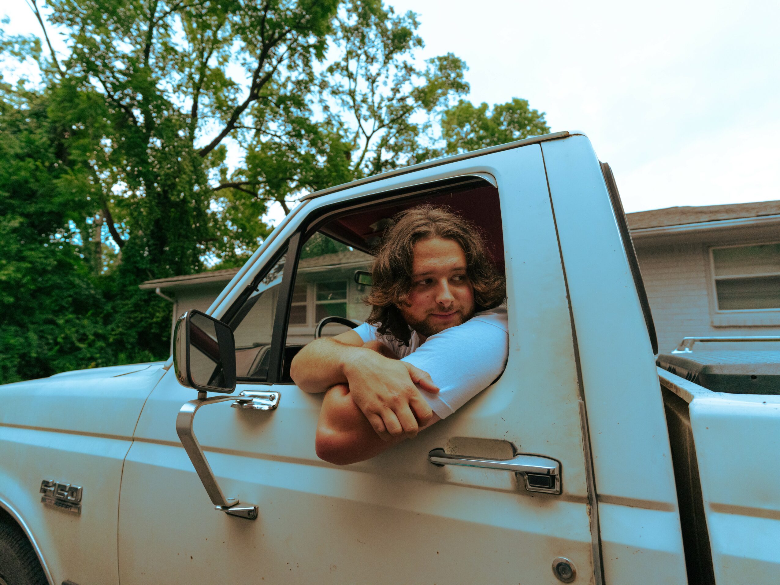 Person with curly hair and glasses leaning out of a pickup truck window, arms crossed, outdoors with trees in background.