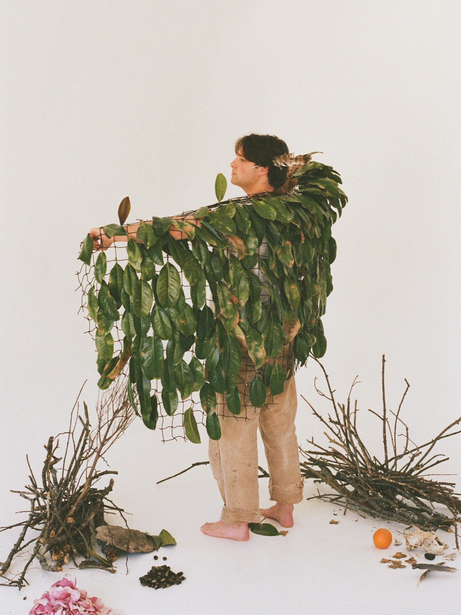 Person standing with a large leafy green cape, surrounded by branches and an orange on the ground, against a plain white background.