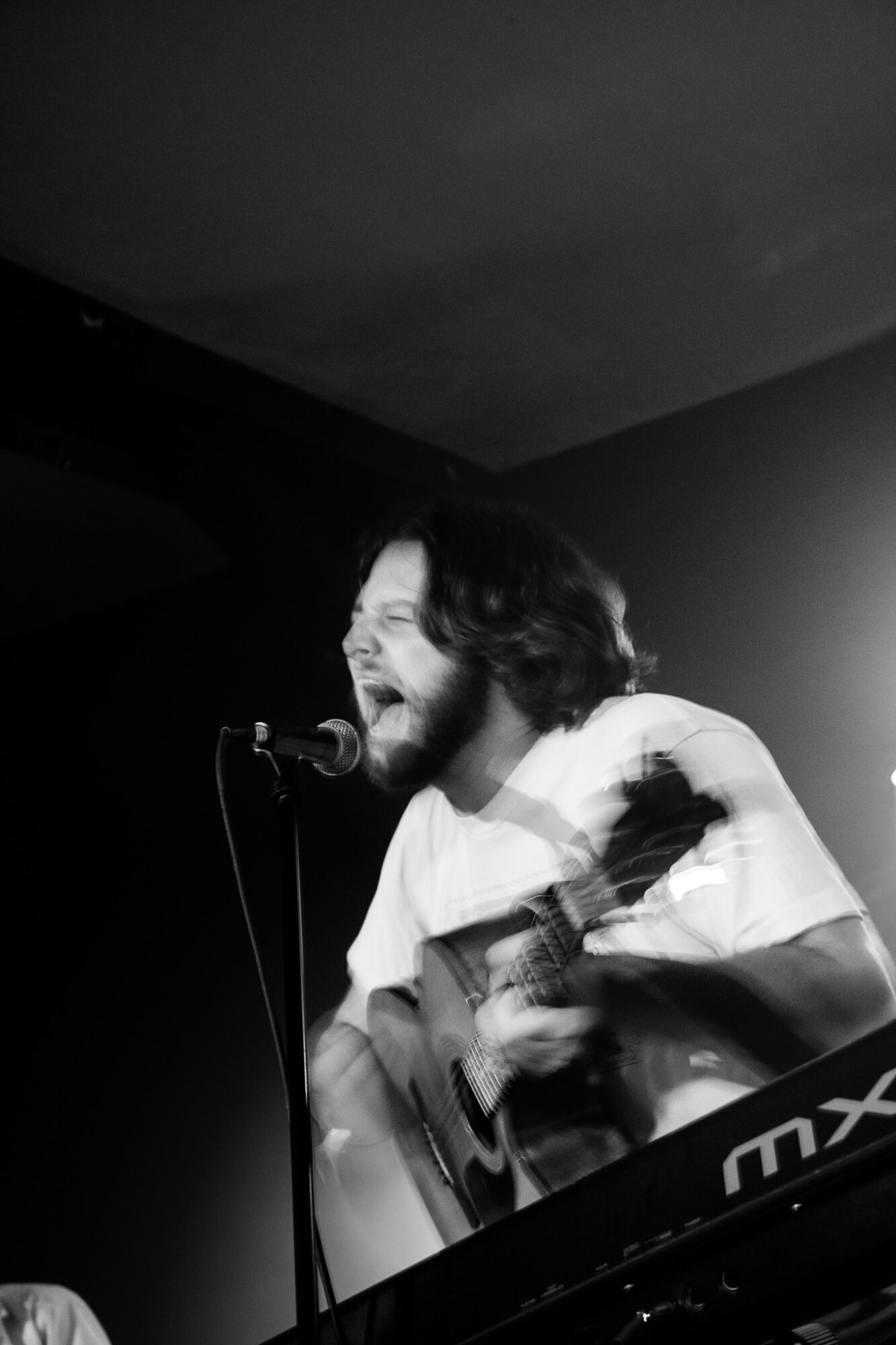 Person singing into microphone while playing guitar, with keyboard in foreground, in black and white.